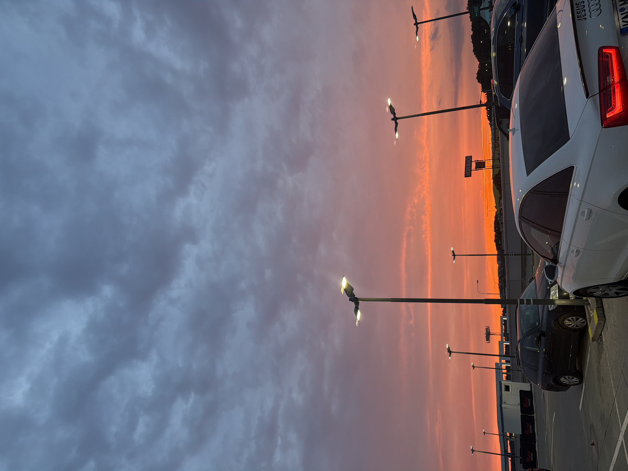 The image captures a dramatic sunset over a parking lot, likely in Prague, Czechia, given the context. The sky is a vibrant canvas of oranges and pinks, transitioning into darker, moody blue and grey clouds on the left side. Several streetlights with illuminated bulbs punctuate the scene, casting a warm glow against the twilight sky. In the foreground, the rear of a white Audi is prominently visible, with its taillights illuminated. Next to it, a dark-colored car is parked. Further in the background, more vehicles and a white portable structure are discernible. A sign with the text "HORNBACH" is visible, hinting at a retail location. The overall atmosphere is serene and picturesque, with the setting sun creating a breathtaking natural spectacle. The time of day is clearly evening, as indicated by the sunset and the lit streetlights.