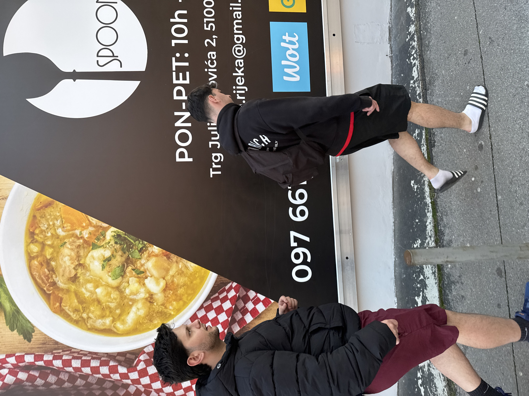 The image depicts two people in front of an advertisement for Spoon, a restaurant. A sign reads "PON-PET: 10h," "Trg Juli," and "ovića 2, 5100," potentially indicating the restaurant's address is on Trg Julija Klovića 2 in Rijeka, Croatia. Also included are "rijeka@gmail." and the phone number "097 66." A bowl of soup is featured on the sign, and the logo for Wolt, a delivery service, is also present. The two people in the image appear to be posing in front of the sign.