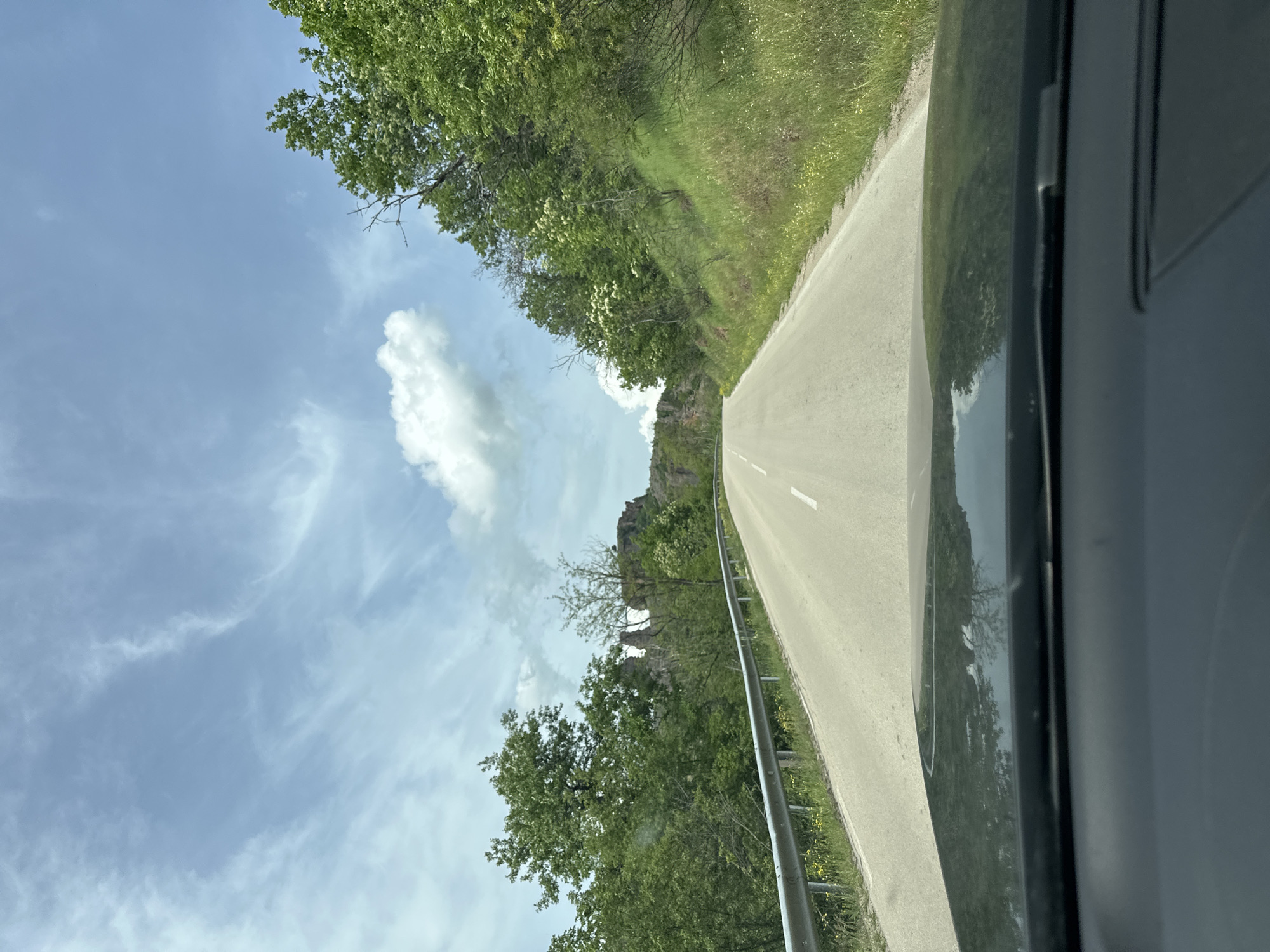 The image depicts a road leading towards the Belogradchik Rocks in Belogradchik, Bulgaria, captured from the perspective of someone inside a vehicle. The road is asphalt, with a white center line visible. The landscape features trees and vegetation along both sides of the road. The sky is blue with scattered clouds. To the left of the road, the Belogradchik Rocks are partially visible. A guardrail is present along the right side of the road. The interior of the vehicle is visible on the right side of the frame. No people are visible.