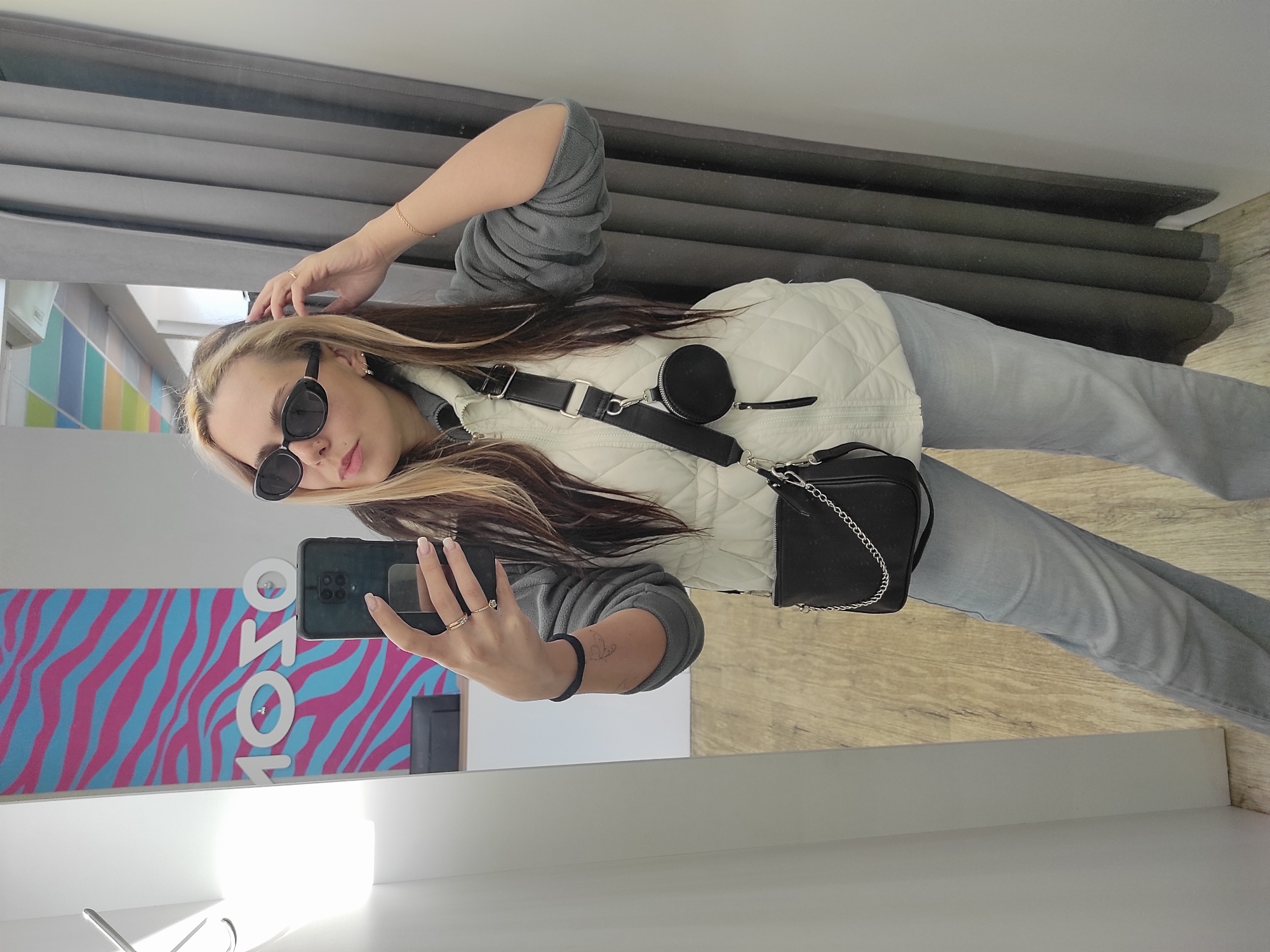 A woman is standing in front of a mirror, taking a selfie with her smartphone. She is wearing sunglasses, a light-colored quilted vest, a gray long-sleeved shirt, and light-wash jeans. She has a black purse with a chain strap slung across her body. Her hair is blonde with dark highlights.
The reflection in the mirror shows a pink and blue zebra-striped design with the word "SOLO" in white letters. The background includes grey curtains. The location is Chaikovsky, Russia.