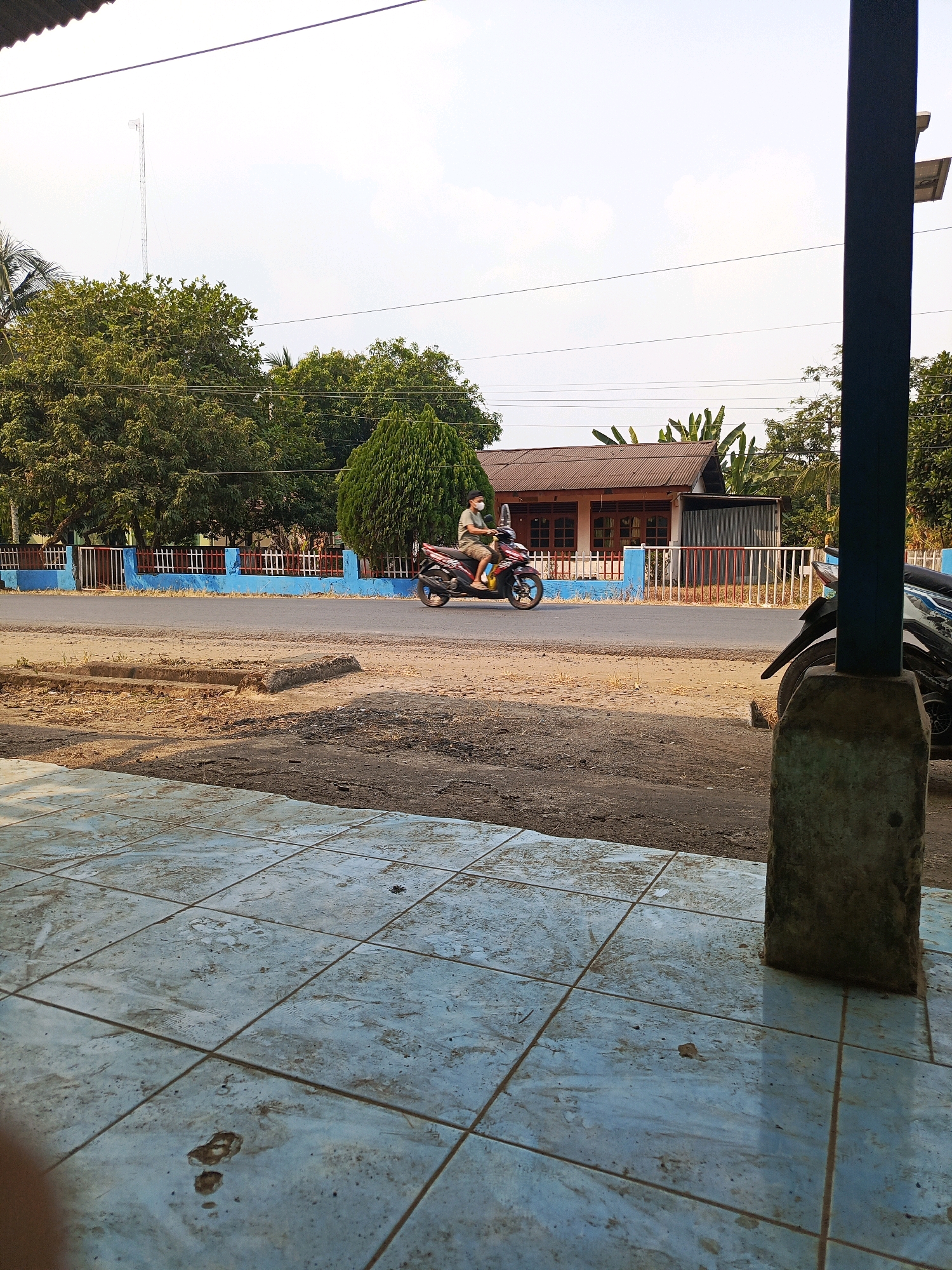 The image captures a street scene in Talang Ubi, Indonesia. In the foreground, a light blue tiled floor is visible, showing signs of wear and dirt. A concrete pillar stands on the right side of the foreground. The midground features a dusty road with a concrete curb. A person on a motorcycle, wearing a face mask and a light-colored shirt with shorts, is traveling on the road. Behind the road, a fence with blue and white sections separates the street from a property with trees and a building. The building has a brown, corrugated metal roof and white walls with windows. In the background, lush green trees dominate the landscape, and a tall radio tower is visible in the far distance against a hazy sky. The overall environment suggests a rural or semi-urban setting.