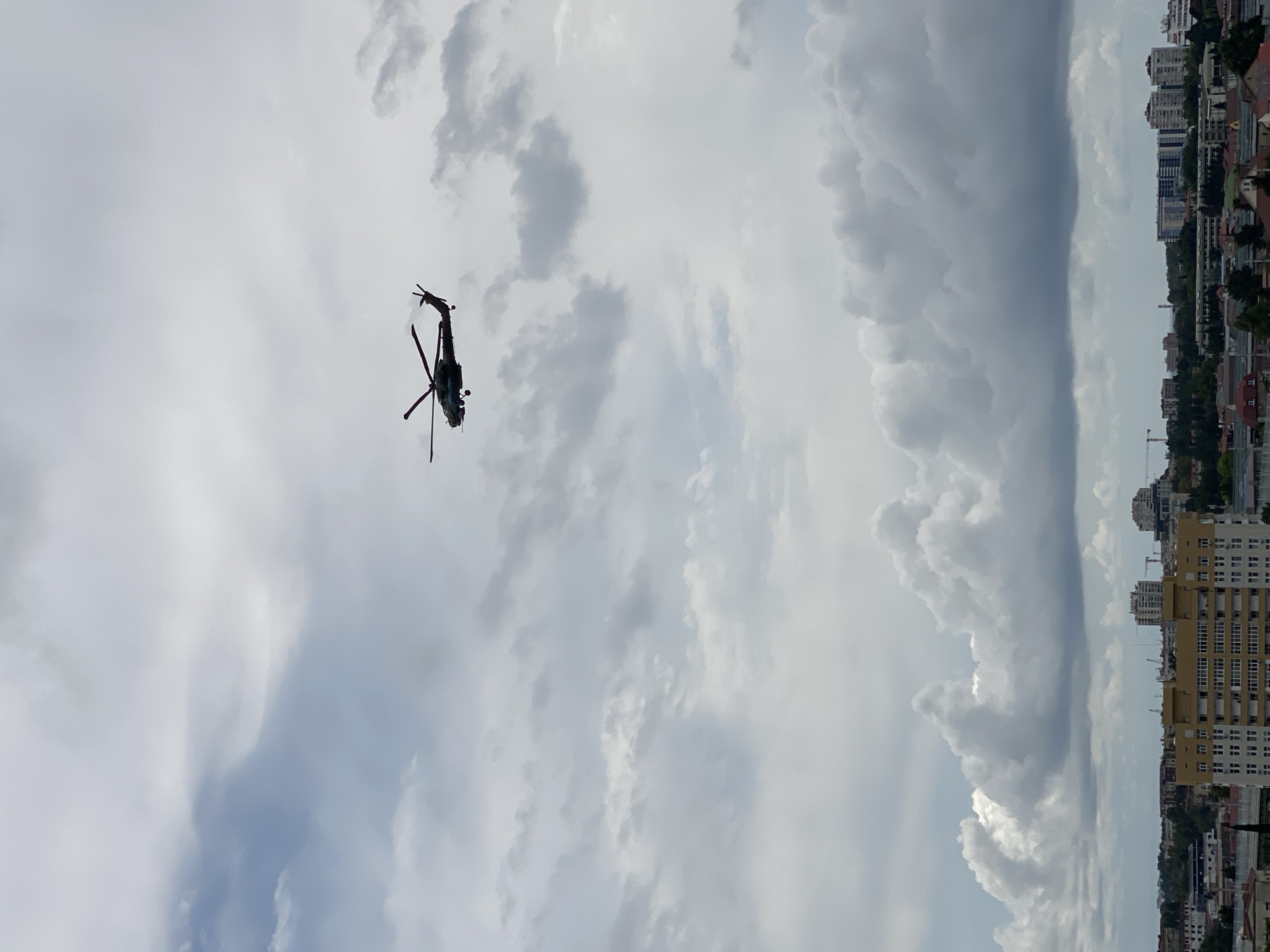 A helicopter is silhouetted against a cloudy sky over a cityscape. The helicopter appears to be a military model with a distinctive tail rotor. Below the helicopter, a dense collection of buildings can be seen, including apartment complexes and what appear to be commercial structures. The sky is filled with various types of clouds, ranging from light, wispy formations to darker, more ominous thunderheads. The overall impression is one of an overcast day in an urban environment. The location is the city of Sochi, Russia.
