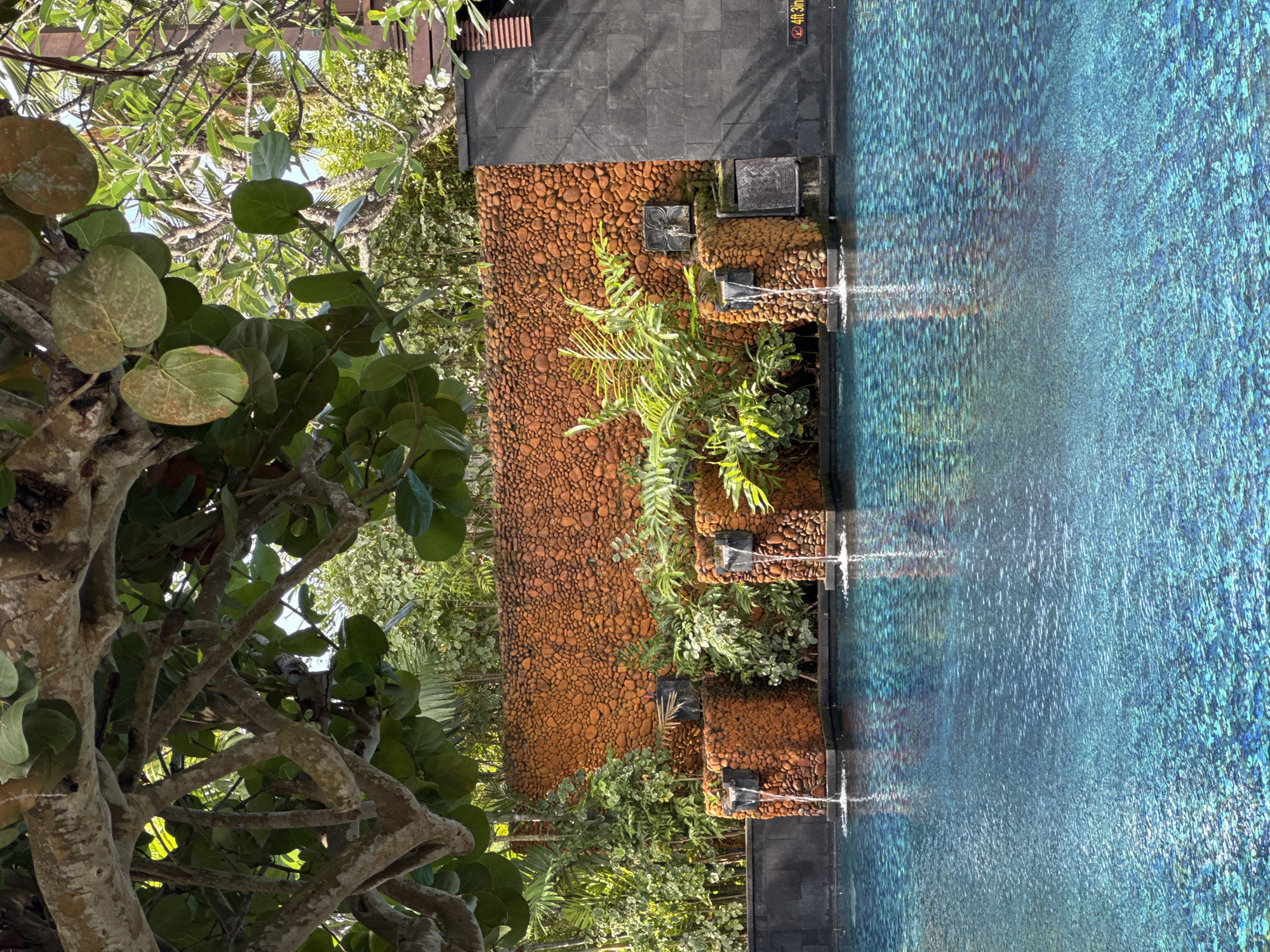 A swimming pool with turquoise water is bordered by a wall of rounded, reddish-orange stones. Four stone pillars, also covered in the same stones, are set against the wall, each with a small waterfall cascading into the pool. Lush green plants are interspersed among the pillars and behind them, adding to the tropical ambiance. A large tree with thick branches and broad green leaves overhangs the scene from the top left. The pool's surface reflects the light, creating a shimmering effect. On the right side of the image, a dark gray tiled wall is visible, with a sign indicating a depth of "4ft 3in". The setting appears to be a resort or hotel in Kuta Selatan, Indonesia, likely an outdoor area designed for relaxation. There are no people visible in the image.