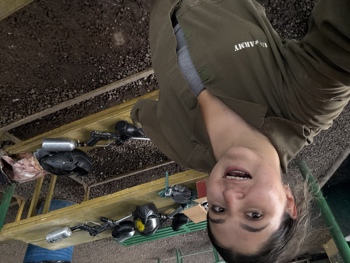 A woman in an olive-green jumpsuit with "U.S. ARMY" emblazoned on the chest takes a selfie. She is indoors with a gravel floor. Behind her, a yellow shelf holds paintball equipment: guns, tanks, and masks. Some of the equipment lies next to a metal rail. The woman looks happy, with slightly parted lips and a smile.