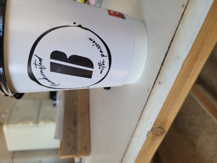 A white disposable coffee cup with a black lid is positioned on a light-colored, possibly white, surface. The cup features a black circular logo containing a bold black letter "B," with the script text "Handcrafted" above and "with passion" below. A small red graphic is partially visible on the right side of the cup. The surface exhibits minor wear and a faint dark line running horizontally. A wooden beam or structural element is visible beneath the surface. In the blurred background, a light-colored appliance, resembling a refrigerator or filing cabinet, is present, along with a wooden tray containing papers. The scene is an indoor environment in Ανδραβίδα, Greece. No people are visible, and no actions or interactions are occurring.