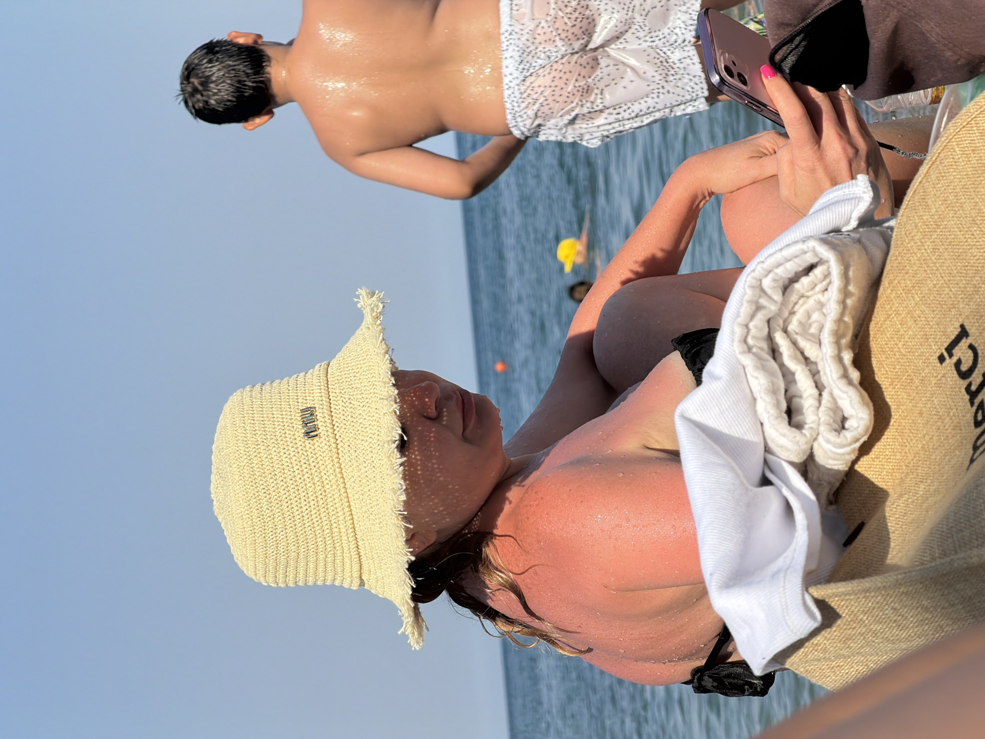 A woman with a straw hat and a black bikini sits on a towel at a beach, holding a pink smartphone. Her skin is red from sunburn. A boy in white patterned shorts stands in the background, his back to the camera. The sea is calm and the sky is clear blue. The scene is likely in Partenit, Ukraine, a coastal resort town.