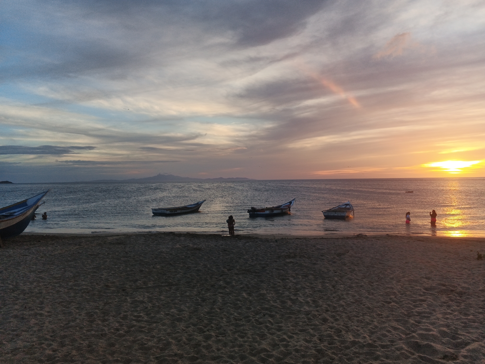 The image depicts a serene beach scene at sunset. The sky is a canvas of soft oranges, pinks, and blues, with wispy clouds scattered across it. A faint rainbow is visible in the upper right quadrant of the sky. The calm ocean stretches to the horizon, where it meets the dusky sky. Several small boats are anchored in the shallow water near the shore. A few people are present on the beach and in the water, silhouetted against the warm glow of the setting sun. The sandy beach in the foreground is textured with ripples, suggesting it is a calm day. The overall atmosphere is peaceful and tranquil.