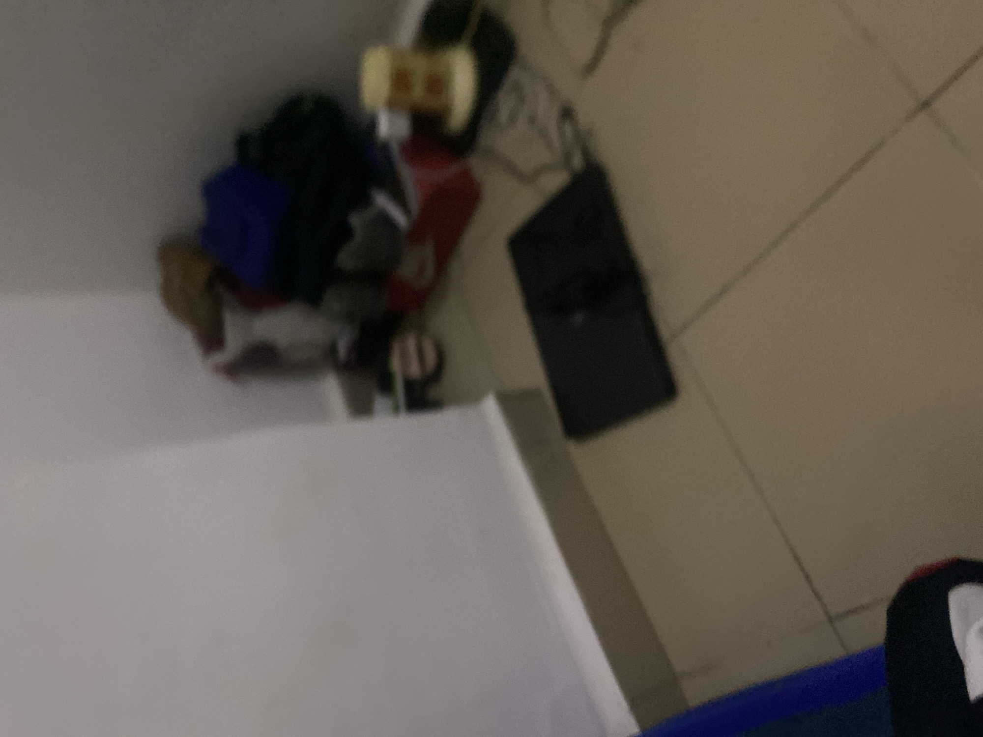 The image depicts a cluttered corner of a room, likely a bedroom or storage area, within a residential dwelling in Howayi, Nigeria. The floor is tiled in a neutral color, with visible grout lines indicating a grid pattern. A white skirting board runs along the base of the wall, which is painted a light, neutral color. In the corner, several objects are piled, including what appear to be clothing items in shades of brown, blue, and dark green, possibly a duffel bag or backpack. A red shoebox is also present, with some white items peeking out. To the right, a cylindrical object with yellow and possibly orange accents sits on a dark surface, possibly a mat or rug. In the foreground, a black, flat object is positioned on the floor, possibly a thin laptop or a similar electronic device. A portion of a blue object is visible on the left side of the image, likely another piece of fabric or an item of furniture. The lighting is dim, creating shadows and a general sense of disarray. There are no people visible in the image.