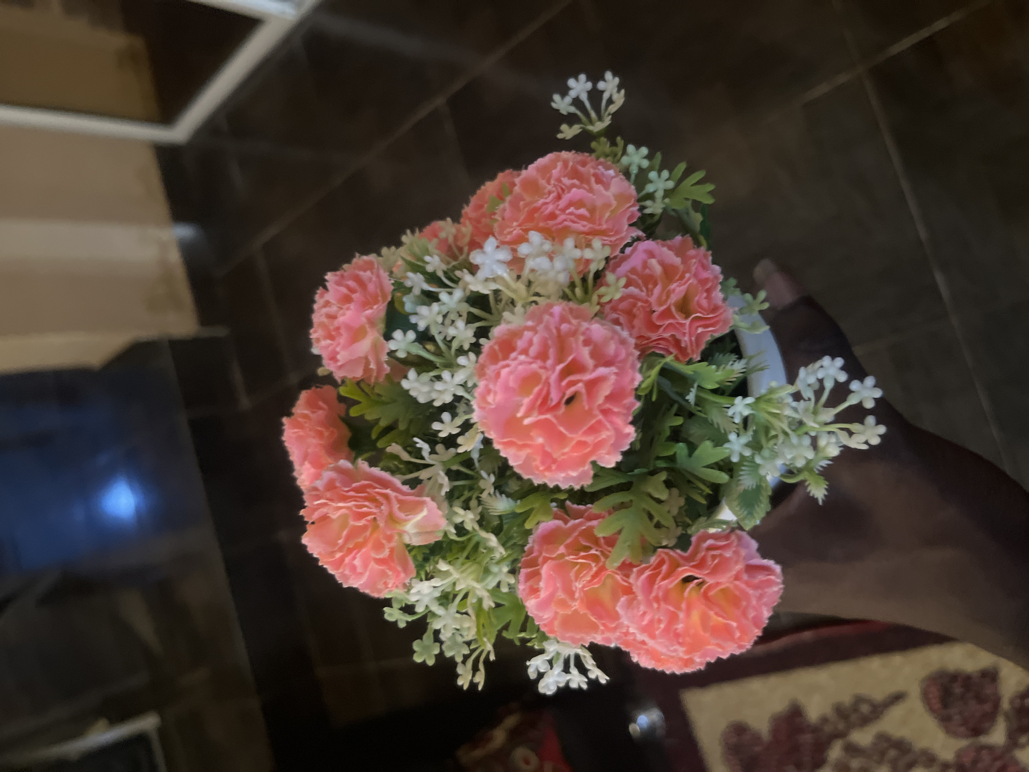 A person's dark-skinned hand is holding a white pot filled with artificial pink carnations and small white flowers, set against a blurred background of a dark tiled floor and a wall. The scene appears to be indoors, possibly in a residential setting within the city of Barakin Akawo, Nigeria, though no specific landmarks or street views are visible.