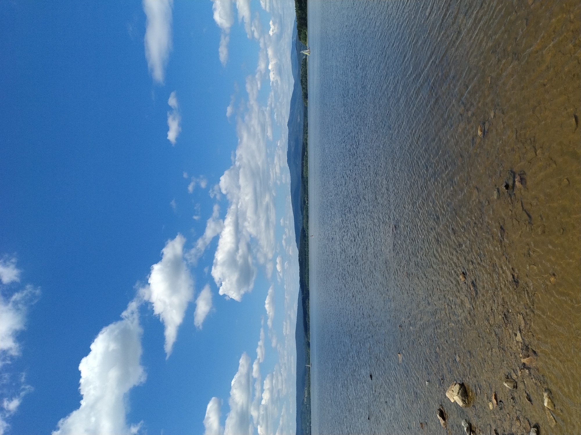 This is a photograph of a large body of water, likely a lake, under a bright blue sky with scattered white clouds. The foreground shows a shallow area with a rocky bottom visible through the clear water. The shoreline is lined with small and medium-sized rocks and pebbles. The main body of the lake is a deep blue-grey, with small ripples on the surface. In the distance, a chain of low, tree-covered hills forms the horizon. Two sailboats are visible on the water; one is closer to the right side of the image and appears to be white with a dark hull, while another is smaller and further to the left. The image captures a serene, natural landscape.