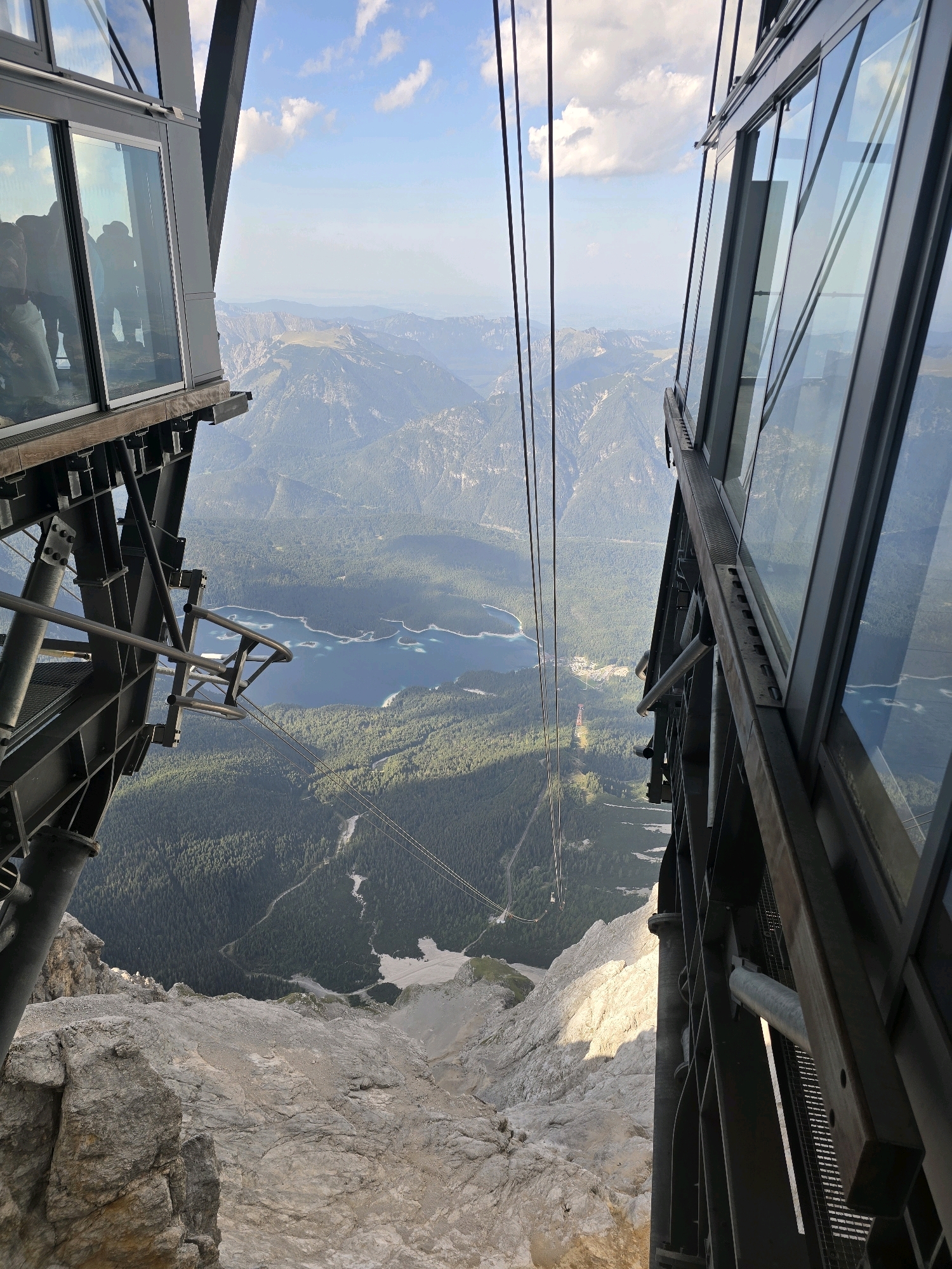 This is a vertical, eye-level, wide shot of a perspective looking down from a cable car station in Grainau, Germany. In the foreground, the metal and glass structures of the cable car station are visible on both sides of the frame. One side features a platform with people on it, their reflections visible in the glass. The other side is a sheer drop, revealing rocky terrain and a distant forest-covered mountain landscape. A bright blue lake, Eibsee, is visible in the middle ground, surrounded by green trees. Several cable car lines are suspended in the air, disappearing into the distance. The sky above is blue with scattered white clouds. The overall impression is of a high-altitude vantage point offering a panoramic view of the Bavarian Alps.