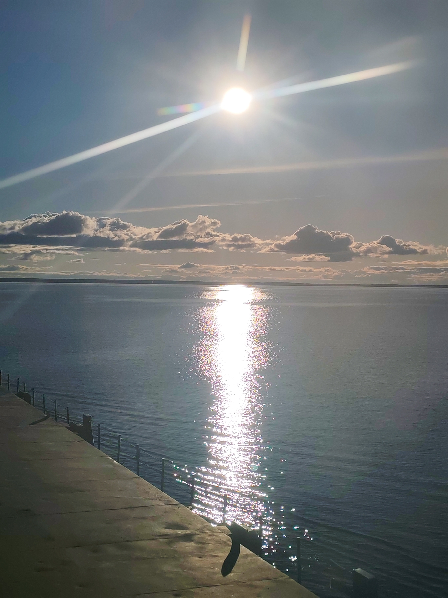 The image depicts a bright, sunny day over a body of water. The sun is high in the sky, producing significant glare and reflection on the water surface. Some clouds are visible on the horizon. In the foreground, there is a concrete walkway or pier with a metal railing. The location is possibly in Kostyaevka, Russia. No people are present.