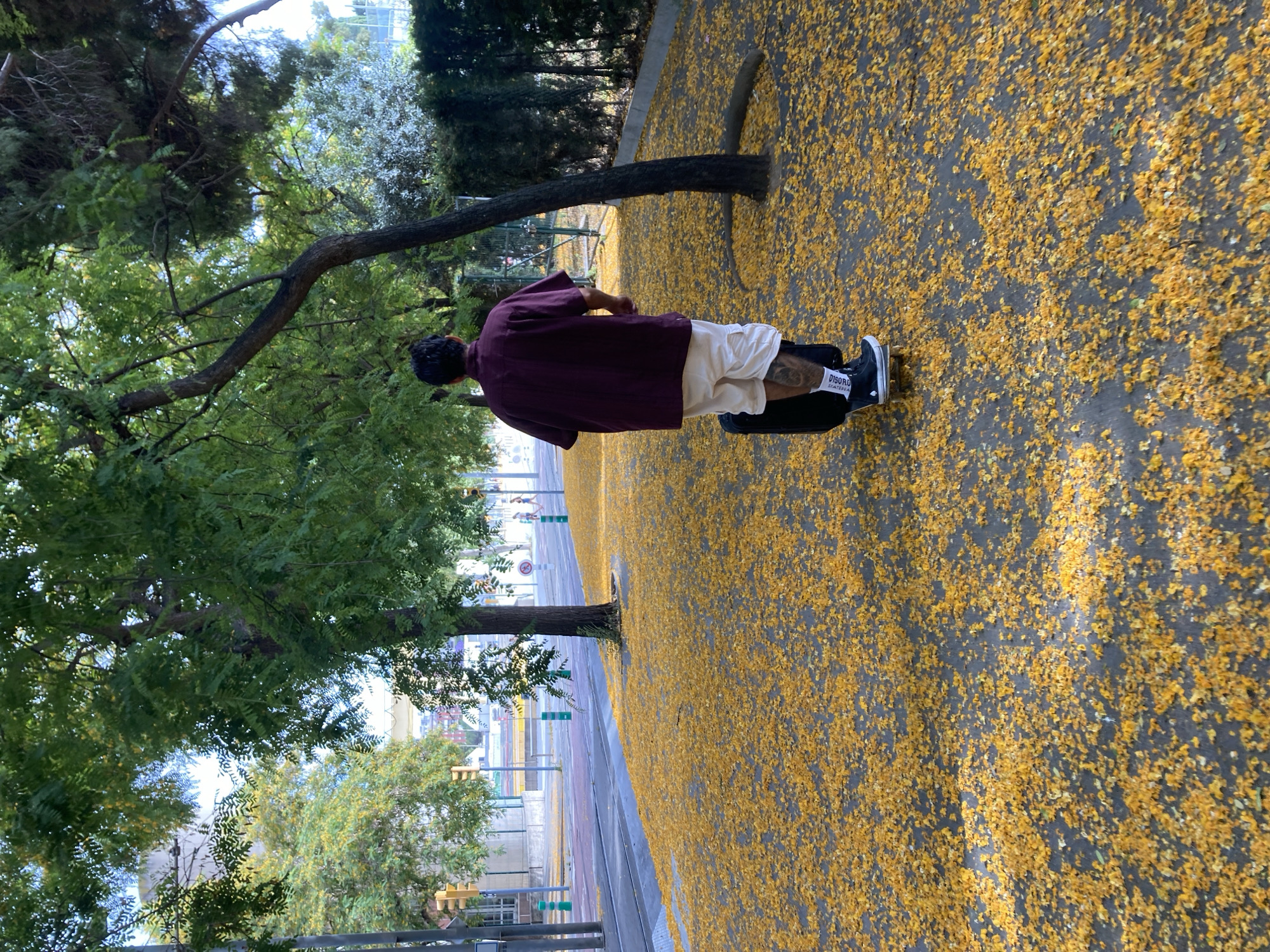 A person is riding an electric unicycle on a pathway covered in fallen yellow leaves. The person is wearing a maroon short-sleeved shirt, white shorts, black socks with white lettering, and black shoes. The unicycle is black. In the background, there is a street with traffic lights and what appear to be tram tracks. Trees with green foliage line the pathway and the street. The scene appears to be in Barcelona, Spain, as indicated by the presence of tram tracks and the overall urban park-like setting.