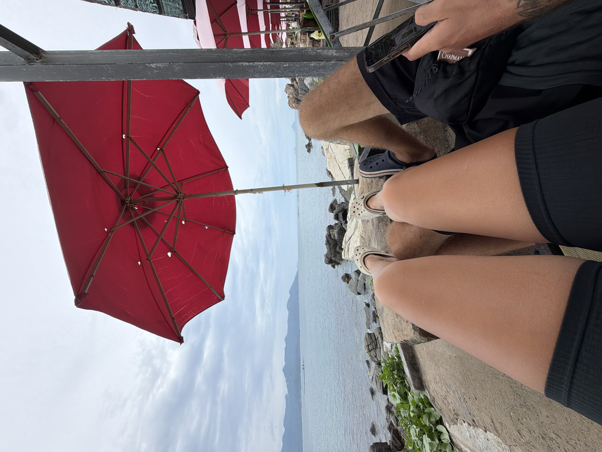 This image captures a relaxed outdoor scene at a waterfront location in Vĩnh Lương, Vietnam, under an overcast daytime sky. The perspective is from a low angle, showcasing the legs of two individuals and the scenic view beyond.
In the foreground, the legs of two people are visible. On the left, a person with hairy legs is wearing black shorts and navy blue Crocs. Their right hand holds a smartphone, and a pack of "CHAPMAN" cigarettes is tucked into a pocket of their shorts. On the right, the smoother legs of another person, clad in black shorts or a skirt and light-colored Crocs, are seen. Both appear to be seated on a concrete or stone ledge.
A large, open red umbrella dominates the left side of the frame, providing shade. Several other red and white-striped umbrellas are visible in the background, suggesting a cafe or restaurant setting. These are supported by a dark metal framework overhead.
Beyond the immediate foreground, a calm body of water stretches towards the horizon, bordered by a rocky shoreline and some green foliage. In the distance, a long range of mountains or hills is silhouetted against the cloudy sky. The overall atmosphere is one of leisure and tranquility, with the individuals enjoying the coastal view.
