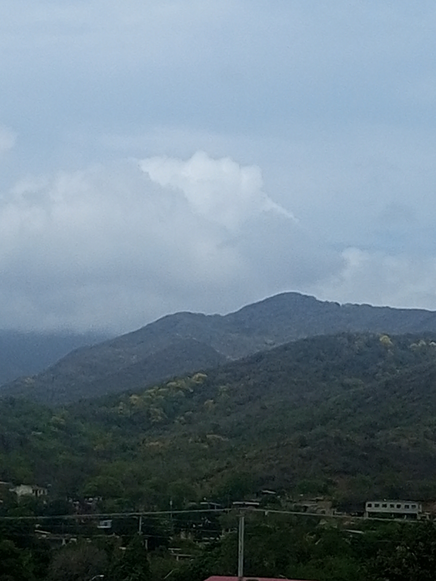 The image depicts a panoramic view of a mountainous landscape under a cloudy sky, with a hint of a settlement in the lower portion. The mountains are covered in lush green vegetation, with some areas showing lighter green patches which could indicate flowering trees or seasonal changes. The sky is overcast with large, diffuse clouds, creating a muted light. In the foreground, at the base of the mountains, a cluster of buildings is visible. One prominent structure on the right is a long, rectangular building with multiple windows. Several other structures and roads are also discernible, suggesting a rural or semi-rural settlement. The overall impression is one of a serene, natural environment with human habitation integrated into the landscape. The scene appears to be situated in the vicinity of La Tagua, Venezuela, a location known for its mountainous terrain and natural beauty.