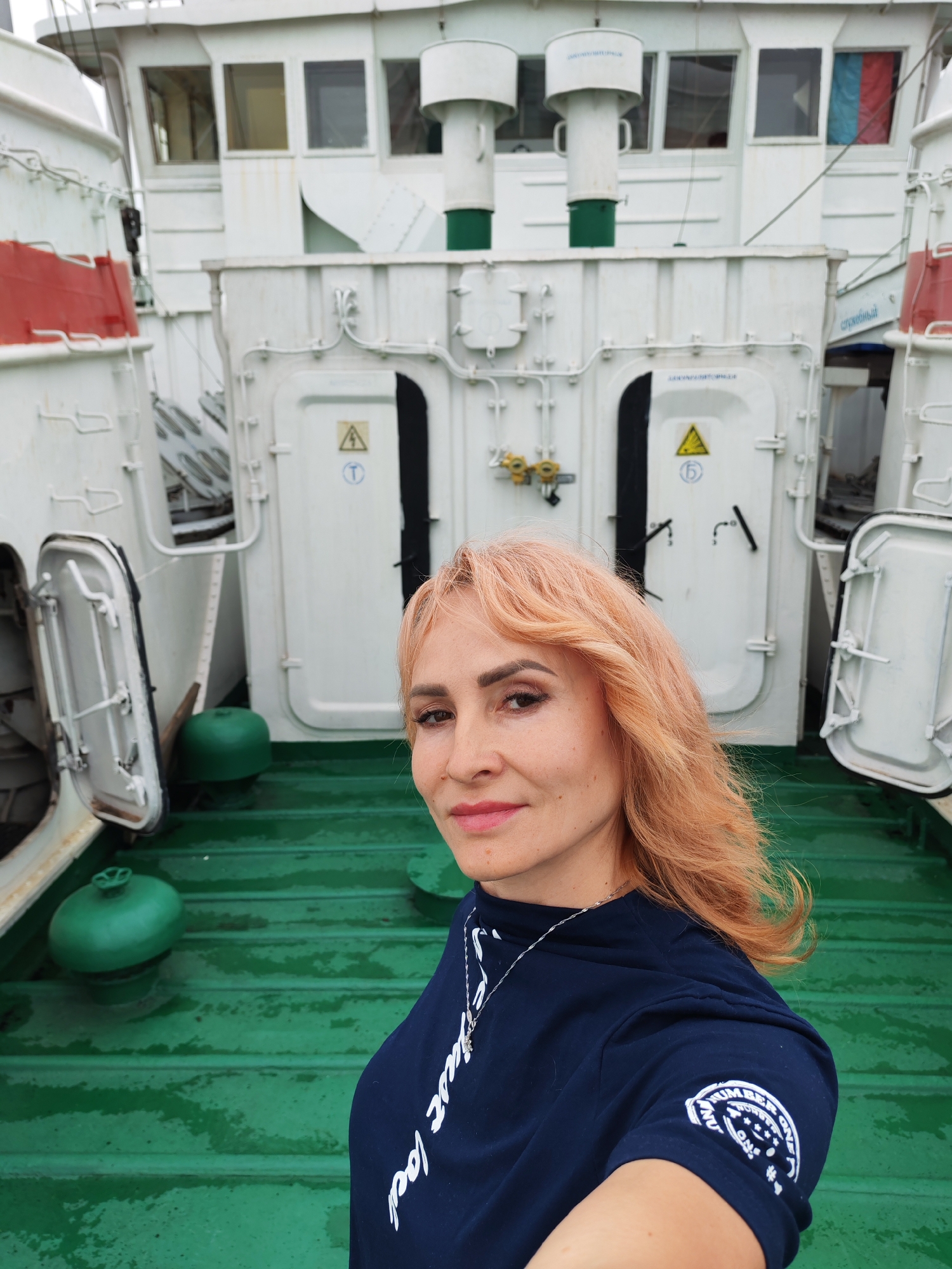 A woman with peachy-blonde hair, wearing a dark blue t-shirt and a silver necklace, takes a selfie on the deck of a ship. The deck is painted a dark green and appears wet. Behind her, the white superstructure of the ship rises, featuring multiple windows, doors, and pipes. Some sections of the ship are marked with red. Two green spherical objects are visible on the deck. The scene appears to be set in Novokemskii, Russia, given the context of the location being provided.