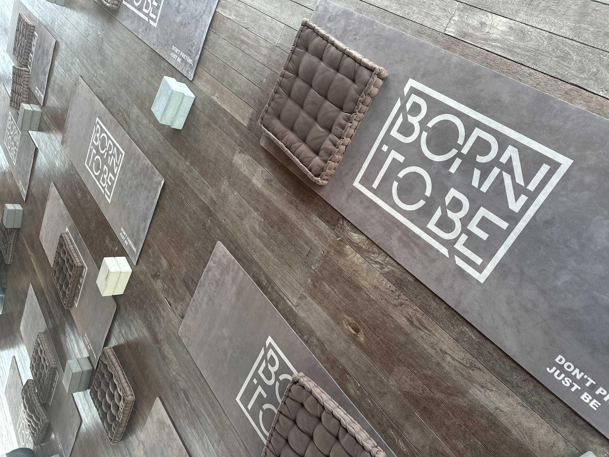 The image shows a yoga studio with multiple yoga mats laid out on a wooden floor. Each mat features a white logo that appears to read "BORN TO BE." Some mats also have white text that says "DON'T PRETEND, JUST BE." Placed on some of the mats are square, tufted brown cushions and stacked grey yoga blocks. The studio is clean and organized, with the mats arranged in rows. The lighting suggests it is daytime, with light coming from above and to the side, creating soft shadows on the floor. The overall impression is one of a peaceful and dedicated space for yoga practice. This scene could be located in the city of Mengwi, Indonesia, known for its cultural heritage and wellness centers.
