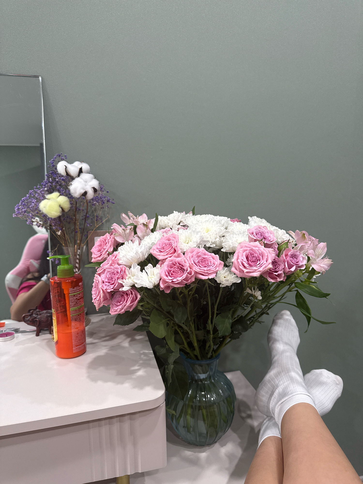 The image depicts a close-up shot of a bedside table or vanity, with a person's legs crossed and extended into the frame, wearing white socks. The setting appears to be indoors, possibly a bedroom. A large bouquet of pink roses, white chrysanthemums, and pink alstroemeria is arranged in a blue glass vase, occupying the central part of the image. To the left of the bouquet, there is a smaller arrangement of dried flowers, cotton bolls, and a yellow puff-like decorative item in a clear vase. Also on the table, an orange bottle with a green pump dispenser is visible, along with a small, dark figurine, possibly an elephant. A portion of a mirror is visible on the far left, reflecting a person in a pink garment. The background wall is a muted, cool green color. No specific landmarks or indicators suggest the location to be Cheboksary, Russia.