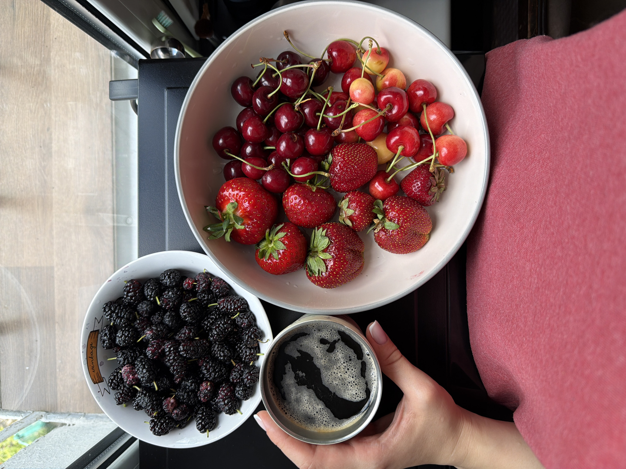 A top-down, medium shot shows a person's hand holding a metal cup of coffee with a foamy top. To the right of the coffee cup is a large, light pink bowl filled with strawberries and cherries. The bowl is placed on a dark surface. To the left of the coffee cup and slightly above it is a white bowl filled with mulberries. The mulberries are dark purple and black. In the background, out of focus, is a window with blinds and a wooden sill. The scene appears to be indoors, possibly near a window with natural light.