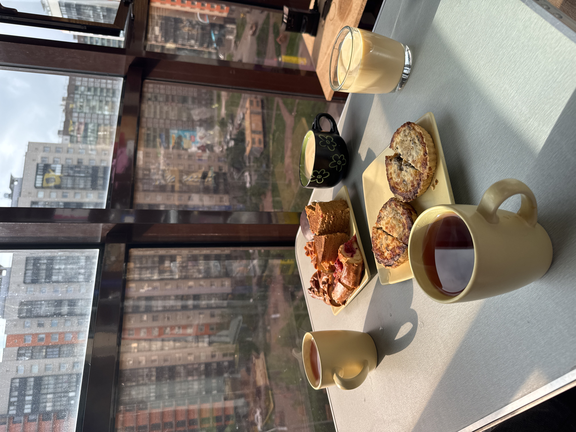 This image captures a breakfast or tea scene in Murino, Russia. The viewpoint is from inside an apartment, overlooking a modern residential area with tall apartment buildings visible through the windows. On a table, various items are arranged: two light-yellow ceramic mugs filled with tea, a black cup with a flower design also containing a beverage, a glass of a light-yellow drink (possibly milk or juice), and two plates with food. One plate has slices of cake, while the other contains halved savory pies or pastries. The overall mood is casual and relaxed.