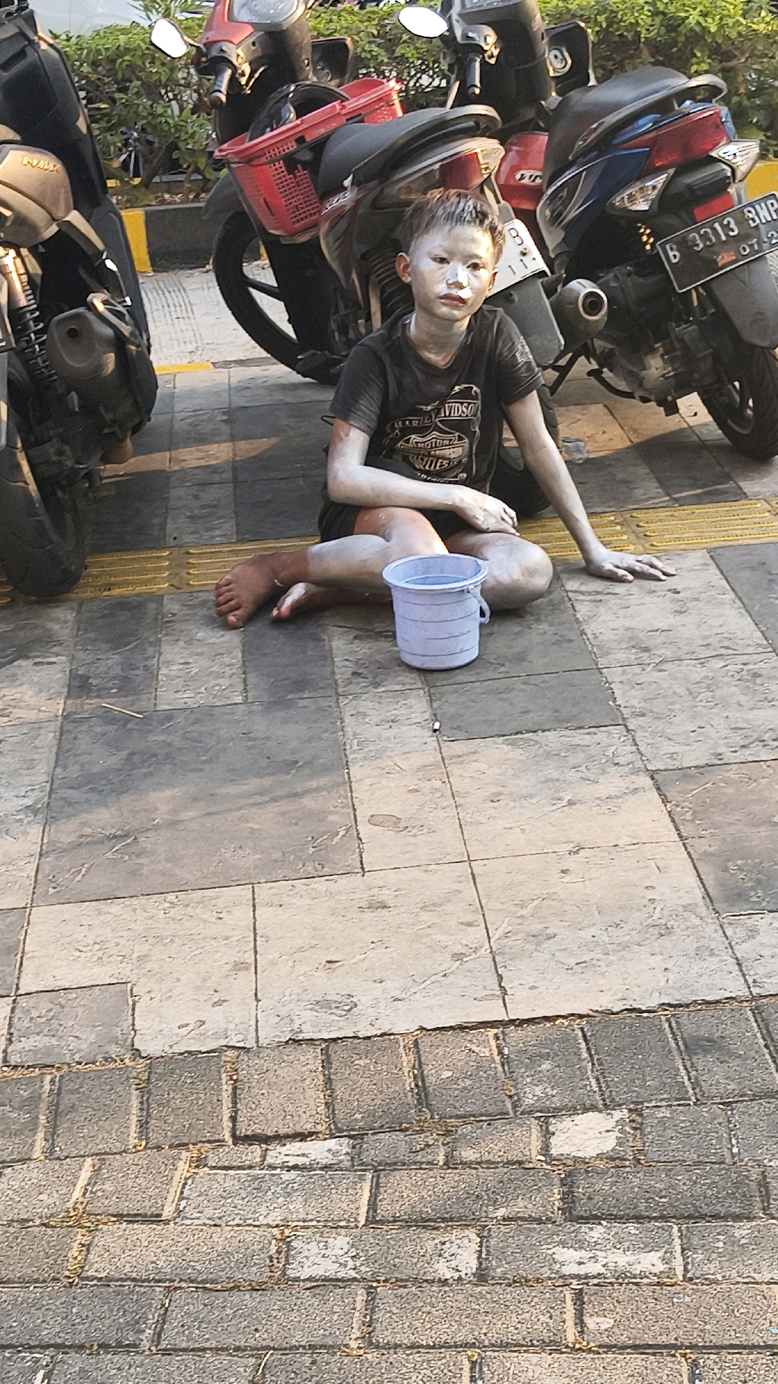 A young person, appearing to be a child or teenager, sits on a tiled sidewalk in West Jakarta, Indonesia. The individual is dressed in a dark t-shirt and shorts. Their face, arms, and legs are covered in a white substance, possibly paint or powder. They are positioned between two parked motorcycles, one red and one blue and black. A small blue plastic bucket sits on the ground next to them, and their right foot is visible, bare. The ground is paved with grey and beige tiles, with a yellow tactile paving strip visible behind the child. The scene is outdoors, with foliage visible in the background.