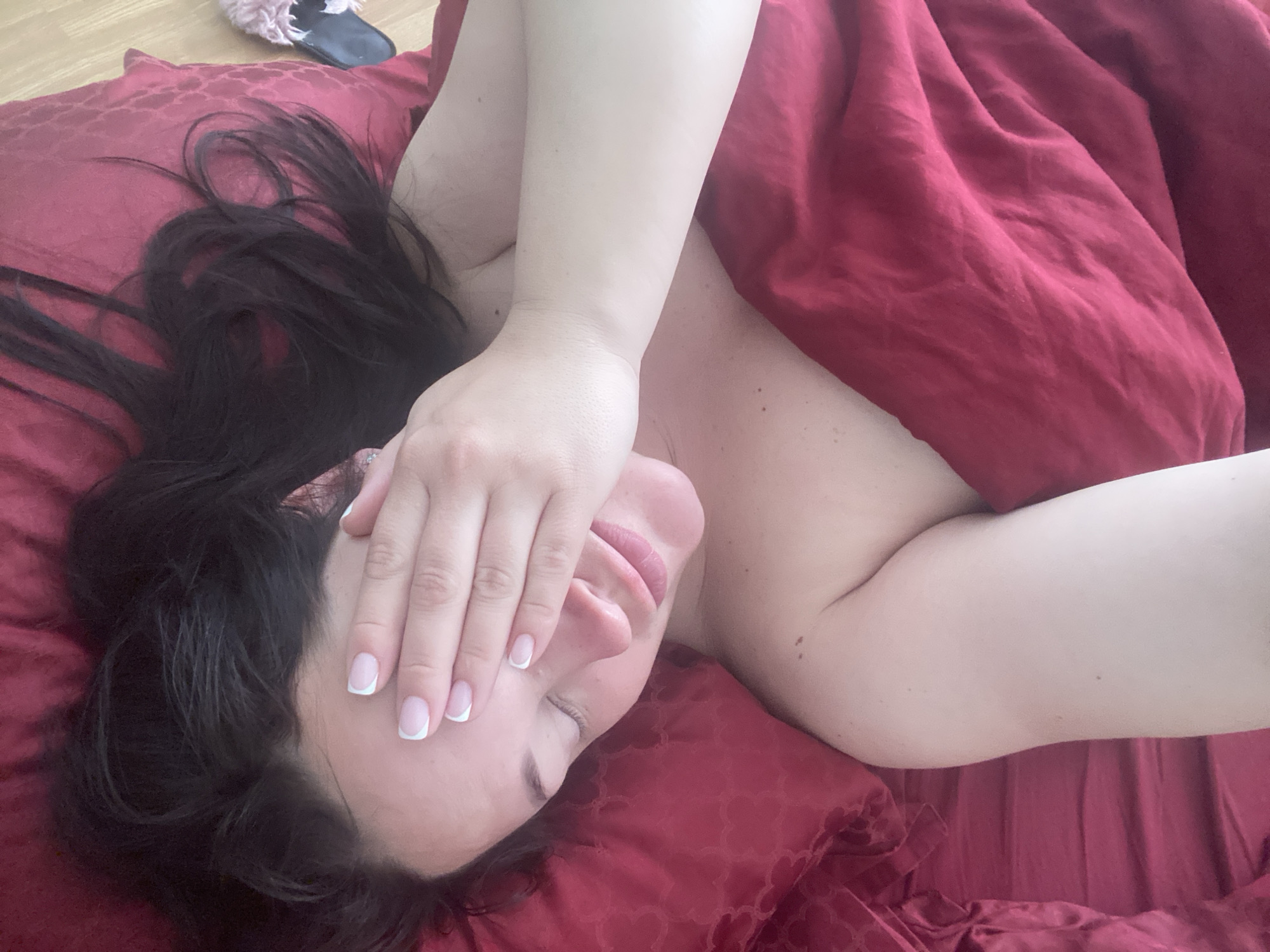 A woman is lying on a bed with red sheets and pillows, covering her face with her hand. Her dark hair is spread across the pillow. Her nails are manicured with a French tip design. A slipper is visible in the upper left corner of the image. There is no indication of the location being Sochi, Russia.