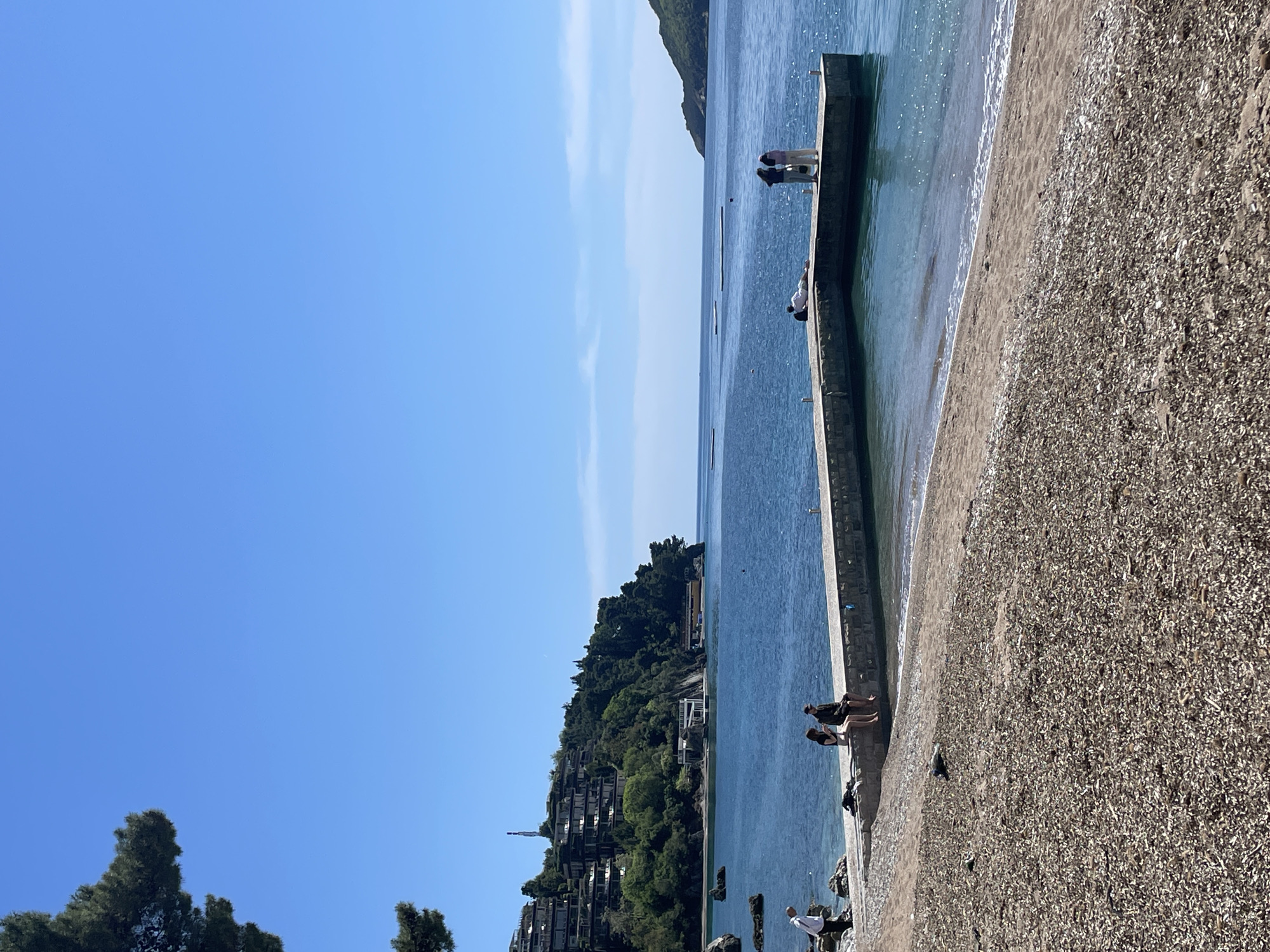 This image captures a bright, serene coastal scene in Budva, Montenegro, on a clear, sunny day. The sky, a vibrant azure blue with a few faint wisps of clouds, dominates the upper left portion of the frame, indicating excellent weather.
A long, concrete pier or seawall extends horizontally across the middle ground, separating the calm, sparkling blue-green water from a narrow, pebbly beach. The water shows gentle ripples and reflections from the sun, with a few dark buoys or markers scattered in the distance.
Several individuals and small groups of people are visible, mostly on the pier or adjacent to it. Towards the far end of the pier, two figures stand together, looking out over the water. Further along, a person in a white shirt leans over the pier's edge, seemingly observing something below. Closer to the foreground, a group of three individuals—two seated on the pier's edge and one standing—appear to be relaxing. Another person in a white top stands at the very end of the pier in the foreground. Their presence suggests leisure and observation of the sea.
To the right of the pier, the beach is composed of small stones and pebbles, stretching along the shoreline. In the background, a lush, verdant hillside rises, densely covered with green trees. Nestled into this natural landscape are several terraced buildings, possibly houses or hotels, with their architecture blending into the hillside and overlooking the bay.
The overall impression is one of tranquility and natural beauty, characteristic of a Mediterranean or Adriatic coastal setting. No visible text is present in the image.