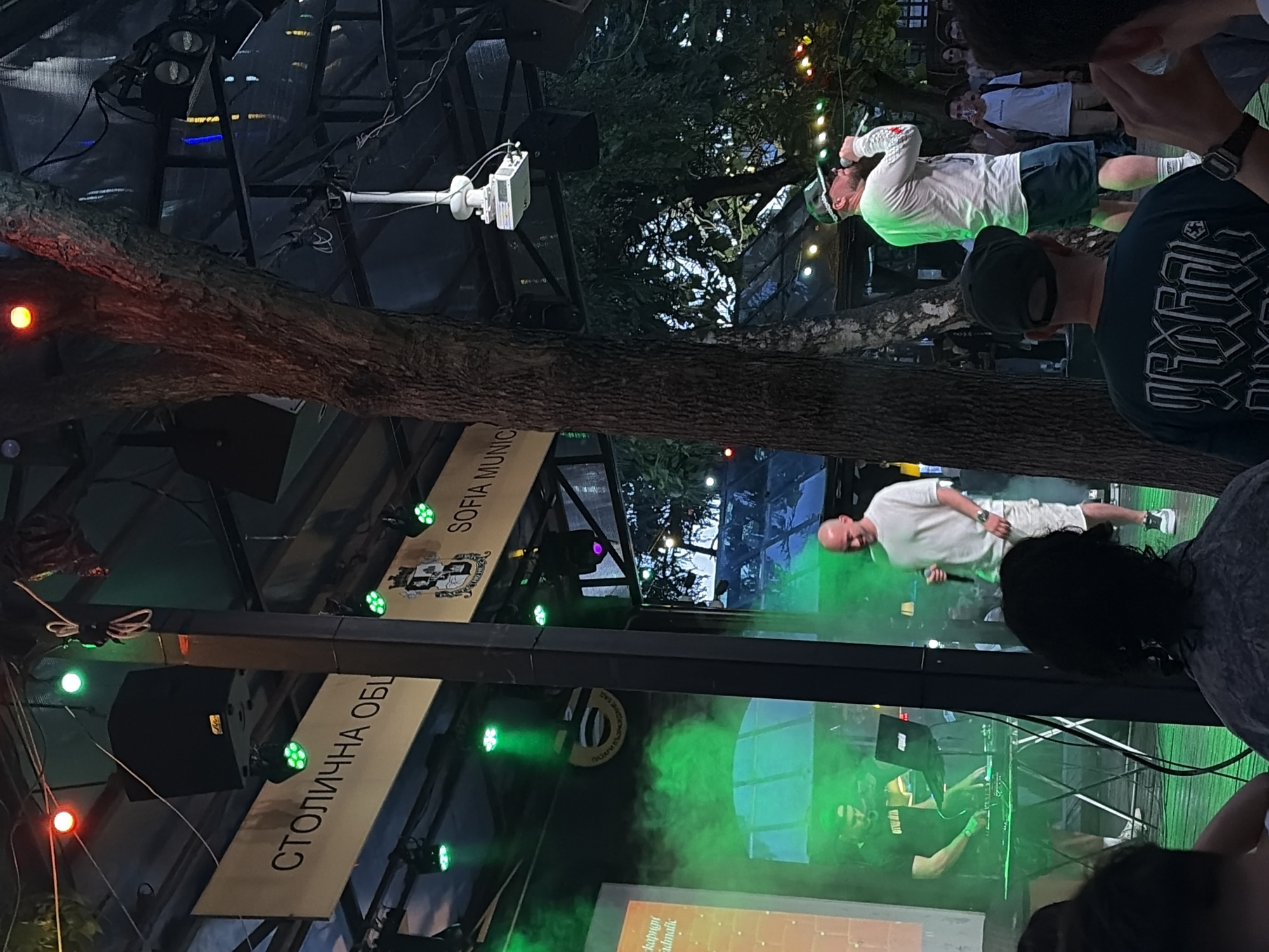 An outdoor event is underway under a covered structure. On the left, a DJ is operating equipment including a laptop and turntables, illuminated by green stage lighting and smoke. In the center, a bald man in a white t-shirt and shorts is holding a microphone and singing or speaking into it. He is lit by green stage lights. Behind him, a large tree trunk is visible. In the background, a banner displays Cyrillic text and "SOFIA MUNIC", suggesting the location is Sofia, Bulgaria. Additional people are visible in the background, some watching the performance, and others further back in the crowd. The scene appears to be a concert or performance.