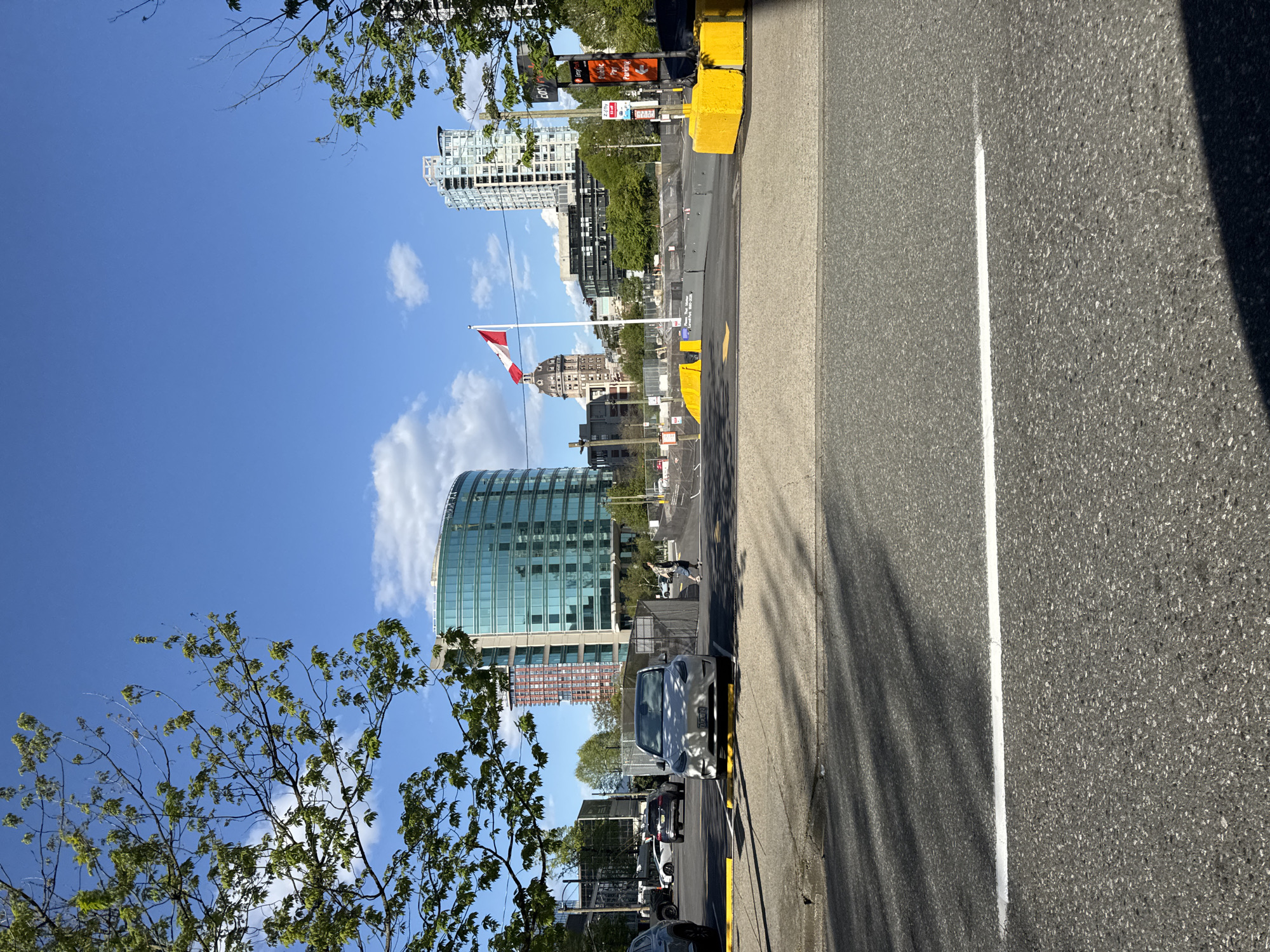 This image captures a street scene in Vancouver, Canada, on a sunny day. Dominating the skyline are modern high-rise buildings, including a curved glass structure. The Canadian flag is visible, suggesting a location near government buildings or a landmark. The foreground features trees with green leaves, typical of the season. The street itself is marked with a white line, and there is a parked car on the side of the road. There are yellow barriers that are likely construction or traffic management tools. The street is lined with parked vehicles. A person walking on the street can be seen.