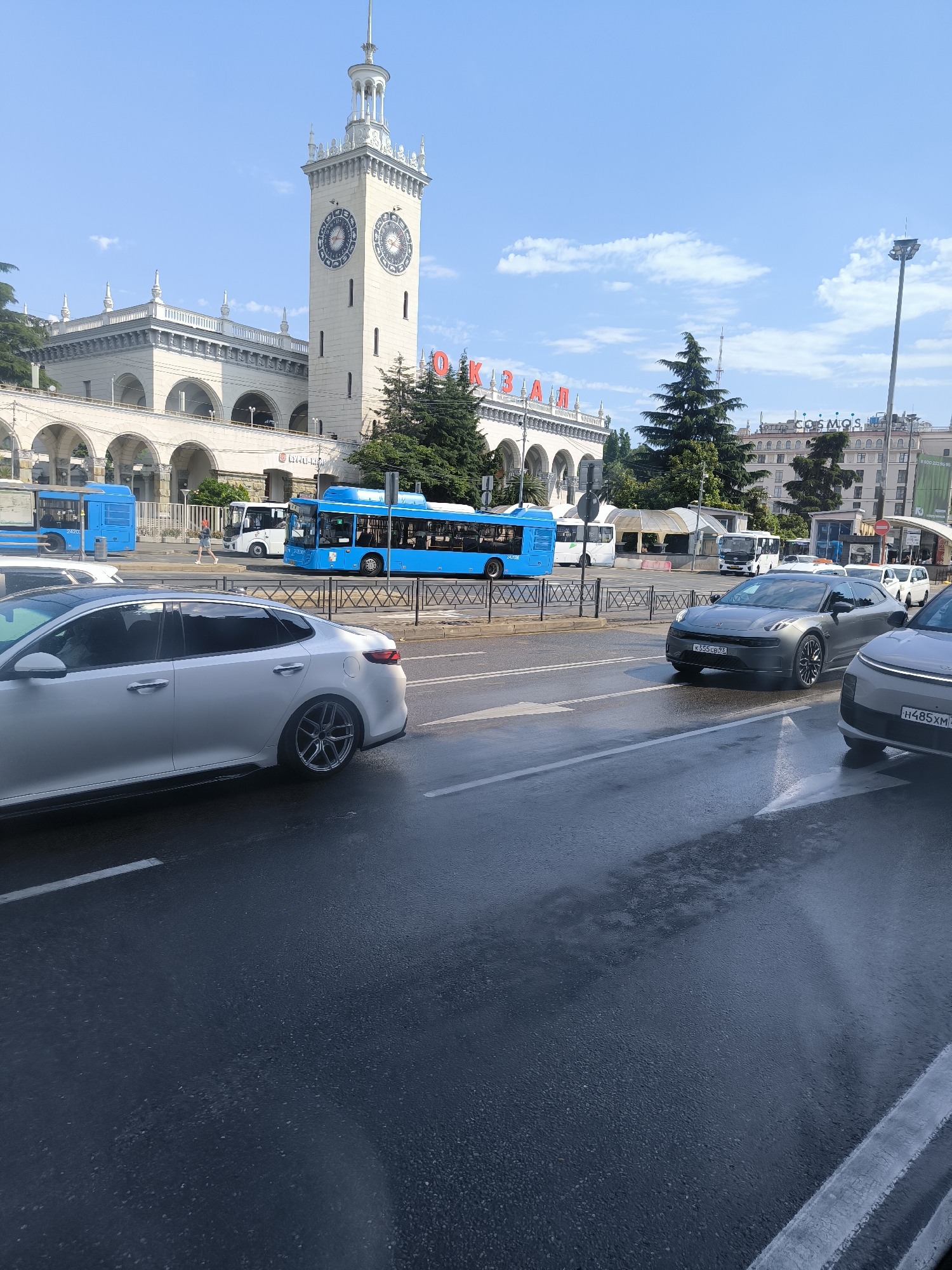 The image depicts a street scene in front of a grand building with a prominent clock tower. The building is light-colored with arched walkways and ornate detailing. Red lettering is visible on the facade of the building, spelling out "ОКЕАН". Several blue buses are present on the road, some of which are in motion. A silver sedan is parked in the foreground, and another car, also silver, is partially visible on the right side. The road appears wet, suggesting recent rain. The sky is blue with some scattered clouds. The overall impression is of a busy urban environment with public transportation and vehicular traffic.