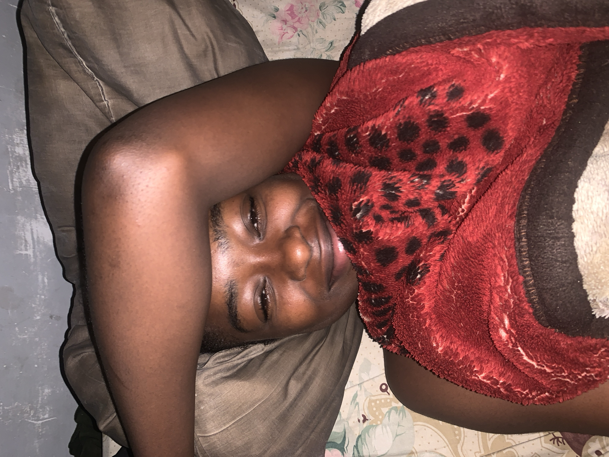 A person with dark skin is lying on a bed. They are positioned on their side, with their arms raised above their head. The person's face is visible, and they appear to be looking directly at the camera. They are covered with a blanket with a red leopard print on it. The bed has a light-colored floral-patterned sheet and is placed next to a dark-colored pillow. The background shows a gray wall. It is nighttime and very dark. There is nothing present to indicate a location in Rafin Par, Nigeria.