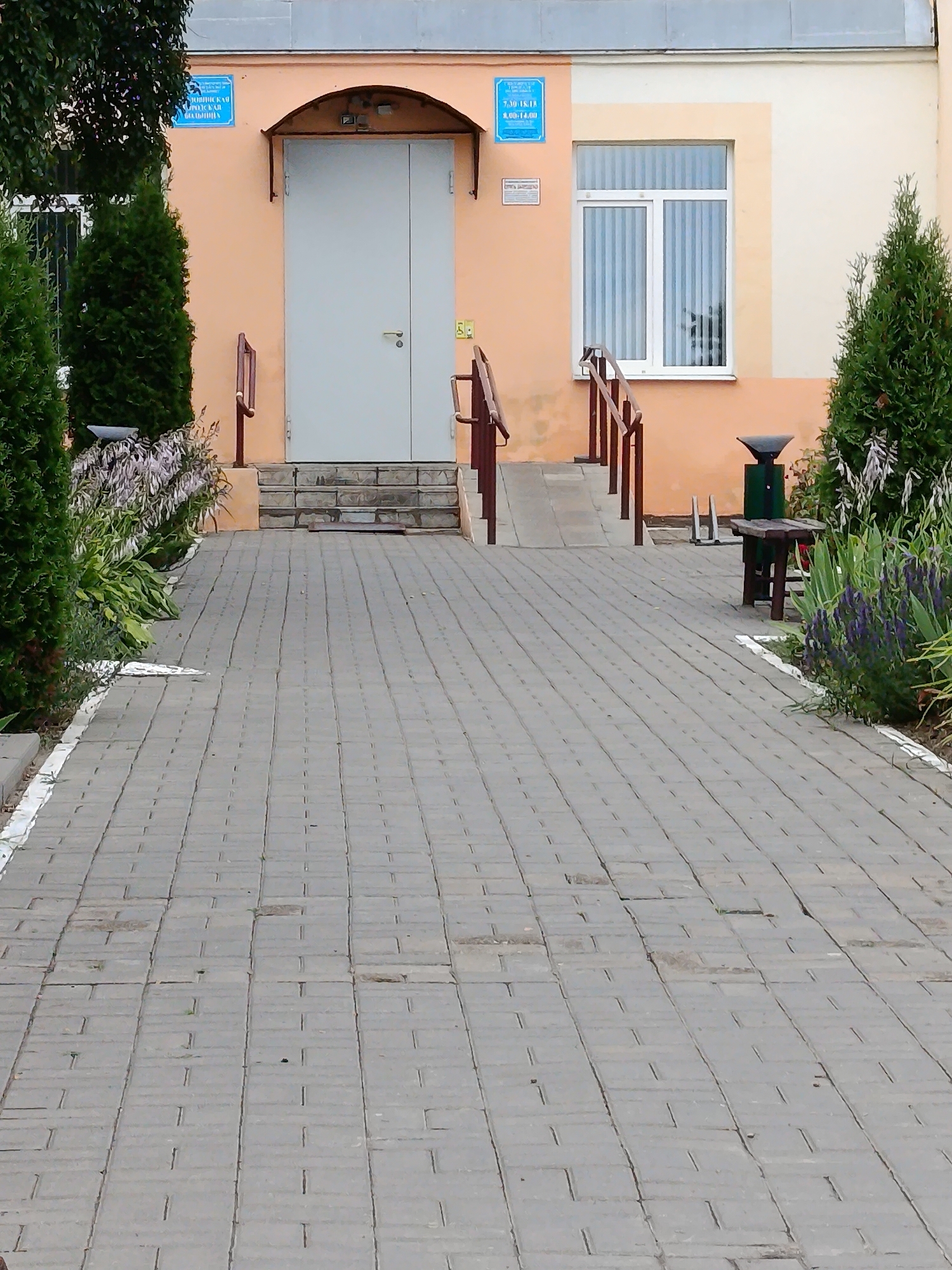 A pathway of gray pavers leads towards the entrance of a building. The building facade is painted in shades of peach and cream. A large, light gray double door is centered on the peach-colored section of the building, with a dark awning above it. To the left of the door, a blue sign is affixed to the wall, displaying Cyrillic text and times. A window with vertical blinds is visible to the right of the door. A ramp with wooden railings runs alongside steps to the left of the entrance. To the right of the ramp, a green trash bin with a wooden bench sits near a cluster of purple flowers and green foliage. Evergreen trees flank the pathway and the entrance. The ground is paved with interlocking gray bricks, with some grass growing through the cracks. The overall scene depicts the exterior of a public building in an outdoor setting.