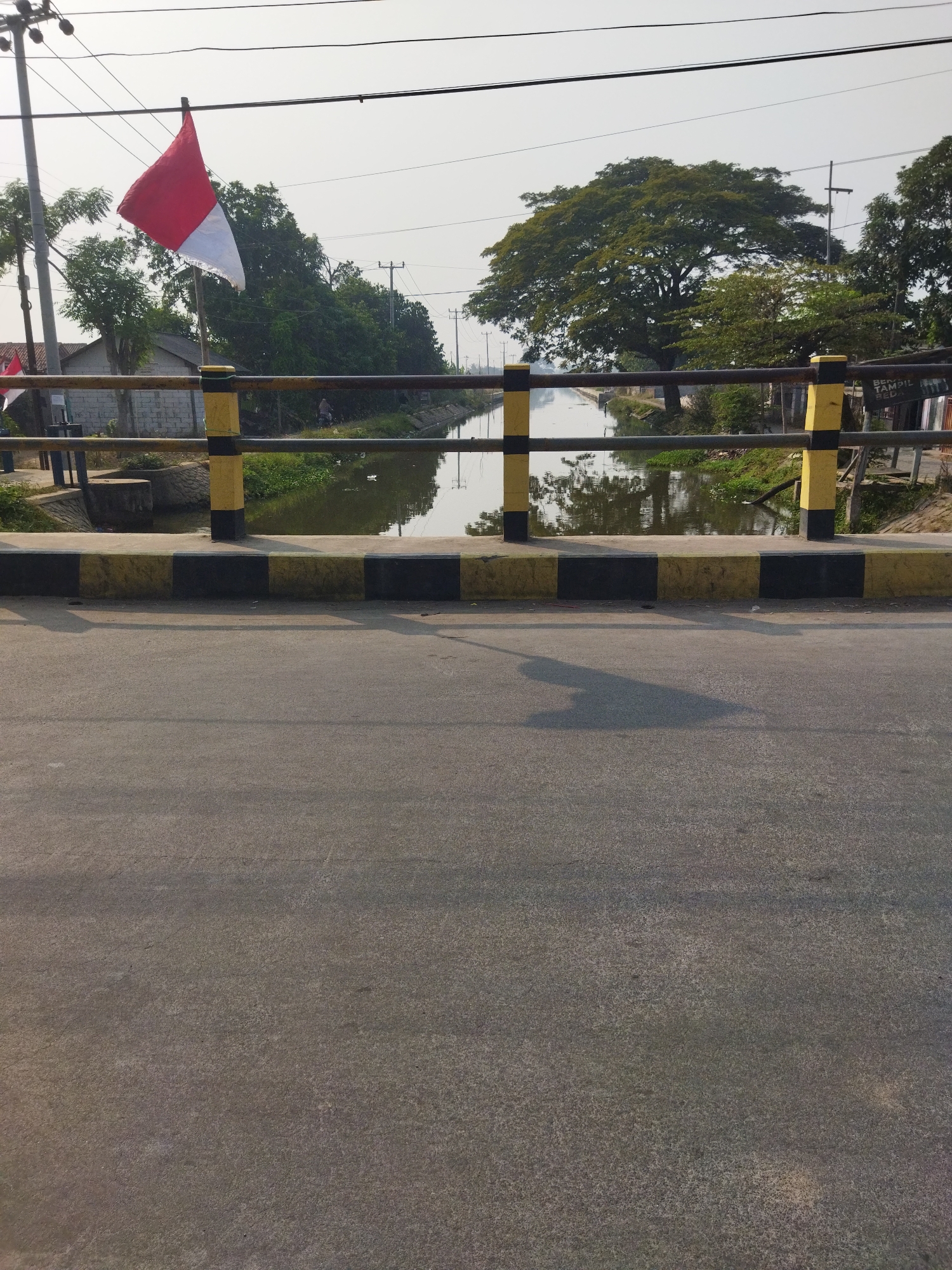 A bridge railing with alternating black and yellow bands overlooks a calm canal in Kayu Bongkok, Indonesia. The Indonesian flag, red and white, flutters from a pole on the left side of the bridge. Trees line the canal banks, and a large, full tree dominates the right side of the scene. The sky is overcast, and the road surface in the foreground shows shadows. A person on a motorcycle can be seen in the distance along the canal.
