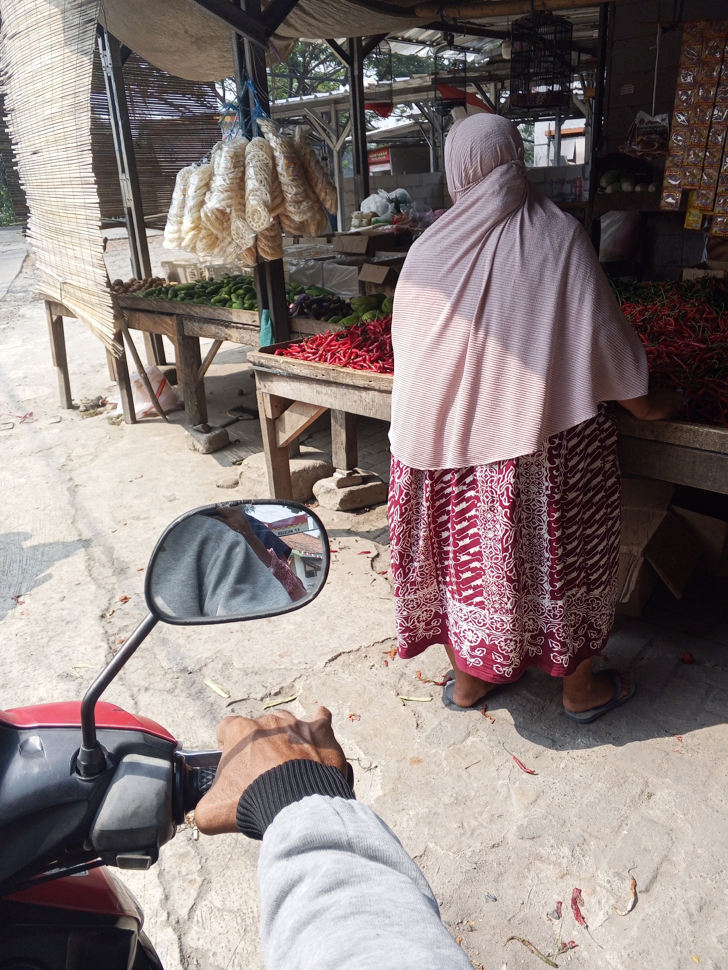 The image captures a street-side market scene in Ciruas, Indonesia, with a focus on a woman standing behind a produce stall. The woman, dressed in a light pink headscarf and a maroon and white patterned skirt, is partially obscured from view as she attends to the goods. In front of her, a large quantity of red chili peppers is displayed on a wooden table. To her left, more vegetables, possibly cucumbers and eggplants, are visible. Hanging above the stall are several bunches of a white, processed food item, likely crackers or crisps. The ground is a rough, concrete surface, with some scattered dried chili peppers and debris. In the foreground, the perspective suggests the image is taken from the viewpoint of someone on a motorbike, with a hand gripping the handlebars and a side-view mirror reflecting a portion of the scene, including the rider's arm and clothing. The overall impression is a vibrant, bustling, everyday market environment in a tropical or subtropical climate.