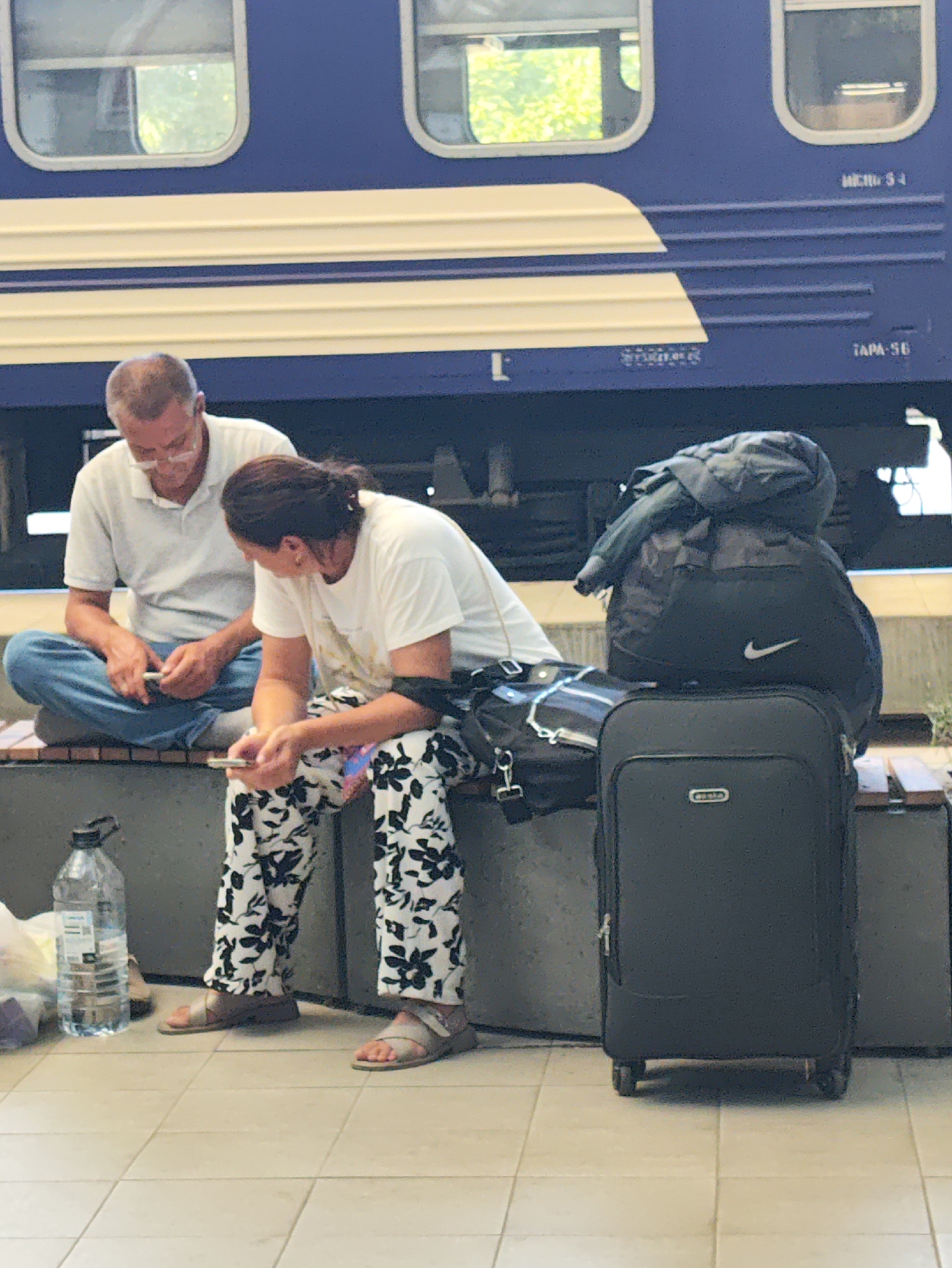 The image depicts a man and a woman sitting on a bench at what appears to be a train station platform. The man, wearing glasses and a white polo shirt, is seated on the left, looking down at his phone. The woman, to his right, is wearing a white t-shirt and patterned pants, also looking at her phone. Between them and to the right of the woman are several pieces of luggage, including a black suitcase with wheels and a backpack on top of it. A large plastic water bottle is visible on the ground to the left of the bench. In the background, the side of a blue train with white trim is visible, featuring several windows. The ground is tiled. Based on the text on the train "TAPA 56", this location is likely the Chișinău railway station in Chișinău, Moldova.