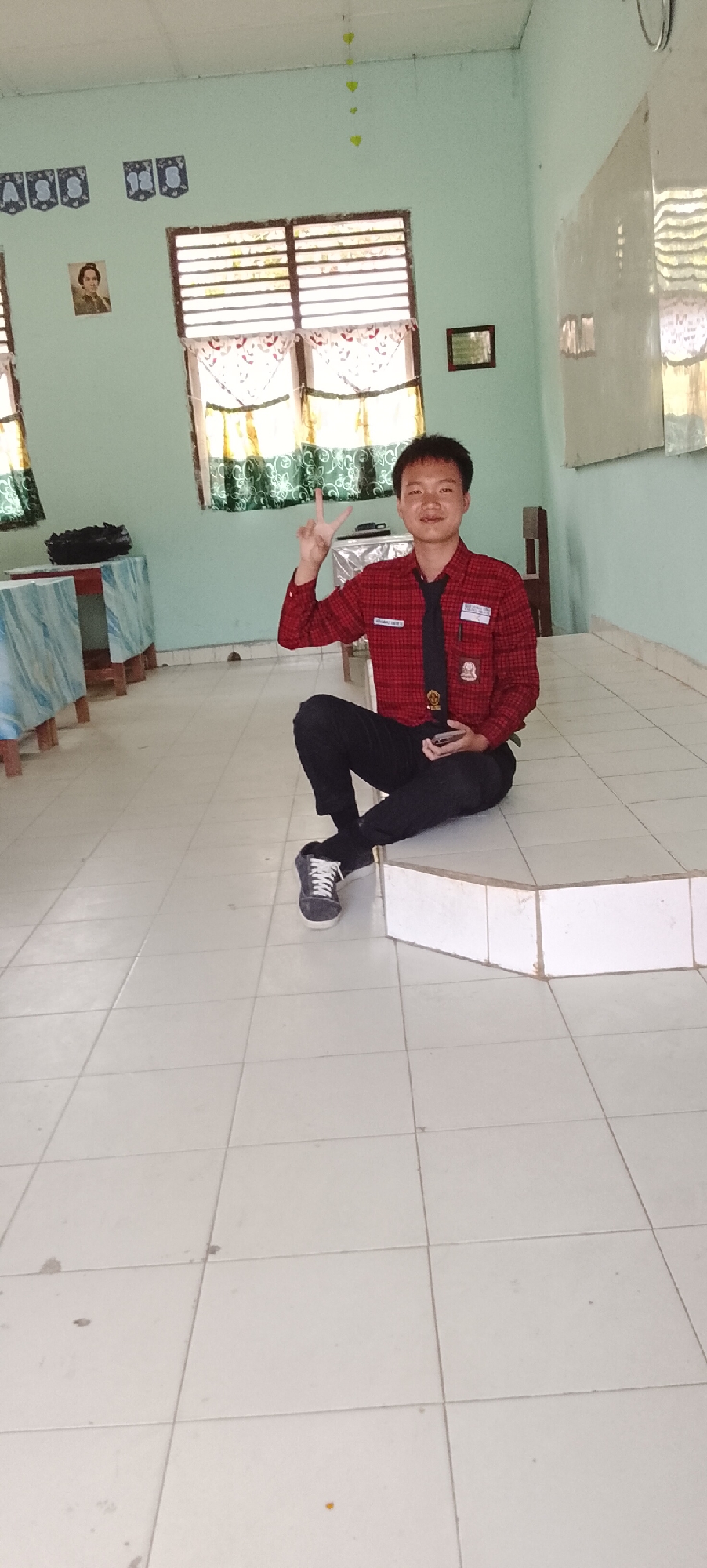 A young East Asian man, wearing a red checkered shirt with a black tie and black pants, is sitting on the floor in a room. He is holding a dark-colored smartphone in his right hand and has his left hand raised with his index and middle fingers forming a peace sign. The room has light green walls and a white tiled floor. There are windows with blinds and curtains on the left side of the room. Two tables with light blue and white patterned coverings are visible on the left. A whiteboard is mounted on the wall to the right. The scene appears to be indoors, potentially a classroom or a communal area, and given the context of Tanjung Setia, Indonesia, it is likely a school setting.