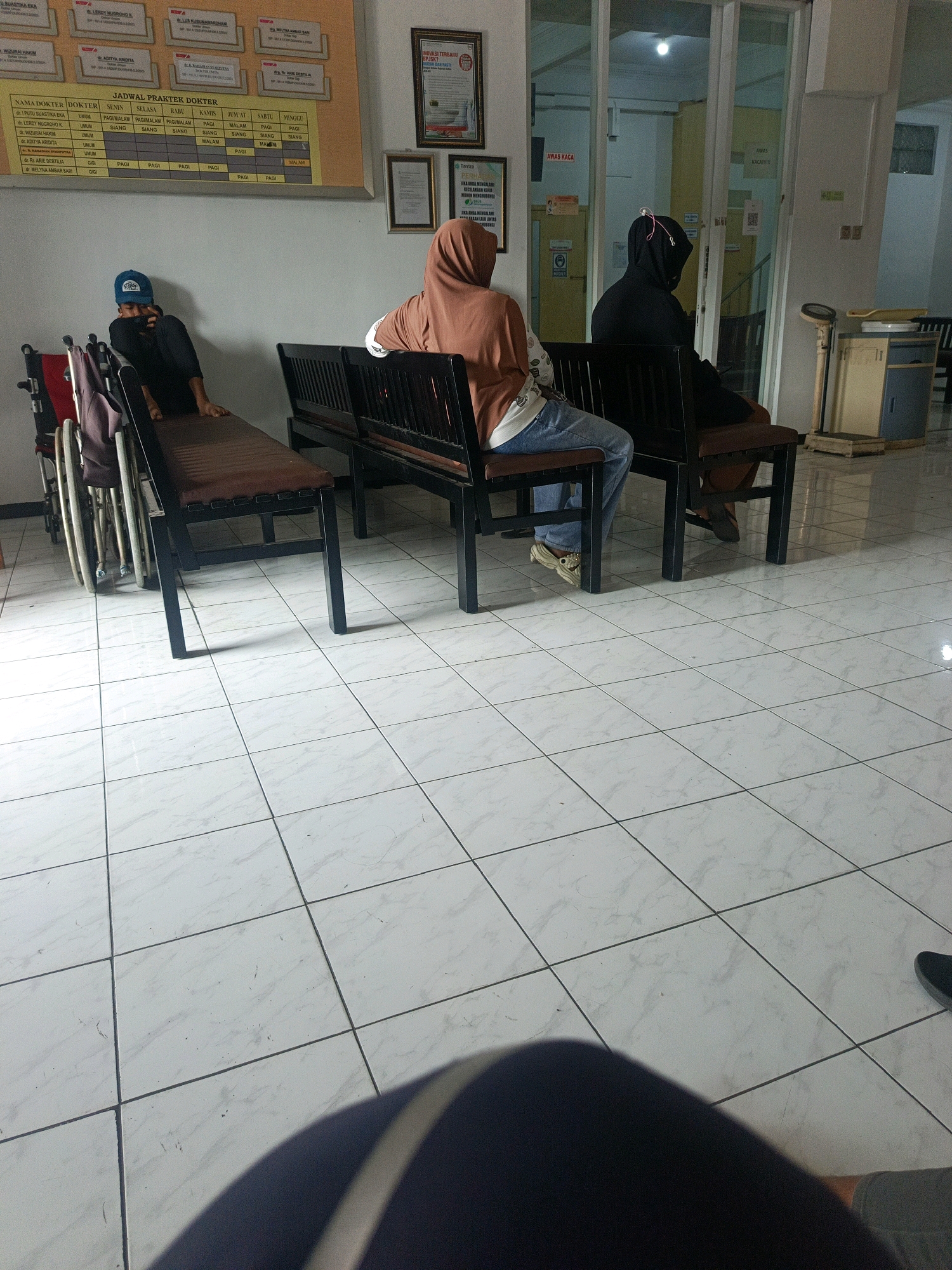 The image depicts the waiting area of a clinic or hospital in Sedati Agung, Indonesia. The floor is tiled with white squares. In the foreground, a person's legs and lap are visible, suggesting they are also a patient or visitor. To the left, a wheelchair is positioned next to a brown bench. A person wearing a blue cap and a dark shirt sits on this bench, leaning back and looking down. Further back, two individuals are seated on a long black bench. One person wears a brown hijab and an orange-brown top with blue jeans, facing away from the camera. The other person is wearing a black hooded garment and also faces away. Behind them, glass doors lead to an interior area with more rooms and signage. To the right of the benches, a medical scale is visible next to a cabinet. A framed document or certificate is mounted on the wall above the benches, and another smaller framed item is below it. A yellow chart with a grid and text is also on the wall to the left. The overall environment suggests a public waiting space within a healthcare facility.