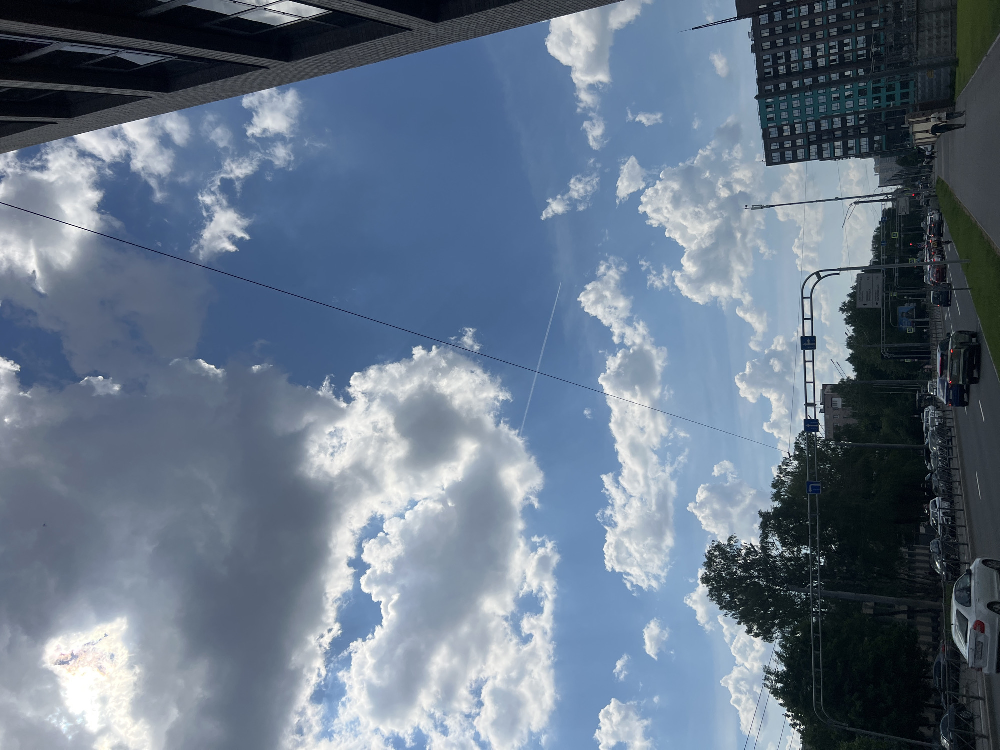 The image depicts an urban landscape under a partly cloudy sky during daylight hours, likely in Saint Petersburg, Russia. The scene is primarily composed of a vibrant blue sky filled with a mix of fluffy white cumulus clouds and darker grey stratocumulus clouds. A bright spot of sunlight pierces through the clouds in the lower left quadrant. A faint white airplane contrail is visible diagonally across the blue expanse.
Overhead utility wires stretch across the sky, originating from the left foreground. In the lower part of the frame, a multi-lane road is visible, bustling with traffic and lined with parked cars on both sides. Lush green trees and foliage flank the road. On the right side of the road, a tall, multi-story building with a distinctive dark and teal-colored facade stands prominent, featuring numerous windows. Further left, another dark-colored building with reflective windows is partially visible, framing the upper left corner of the sky.
Several road signs are scattered along the street. One sign clearly shows a white arrow pointing right on a blue rectangular background, indicating a mandatory direction or turn. Another larger sign structure includes multiple blue squares with white arrows, likely directing traffic for different lanes or destinations. A pedestrian is seen walking along the sidewalk next to the road, near the teal-colored building. The overall impression is that of a typical, busy city street on a clear and sunny day.