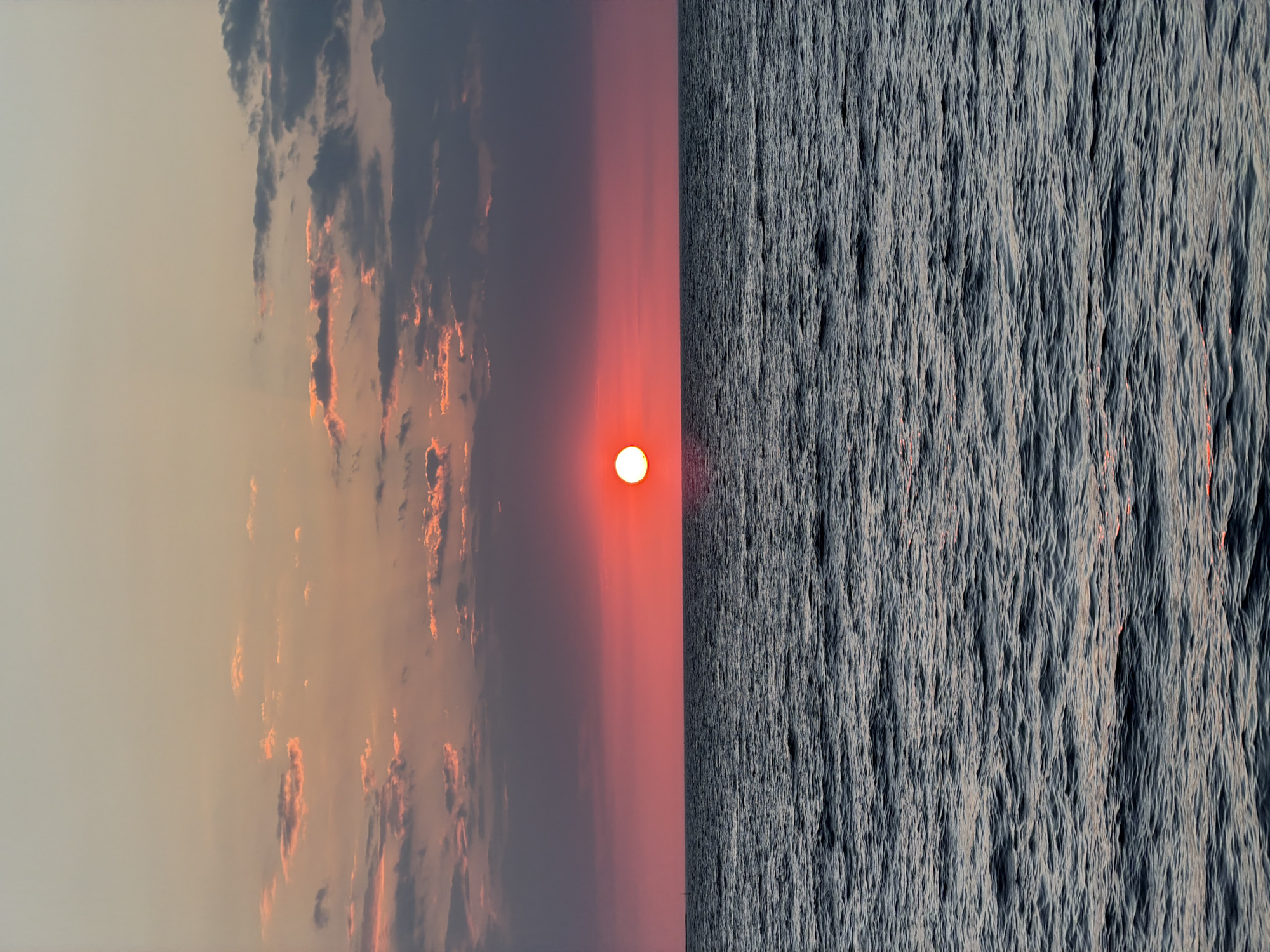 The image presents a split view of a sunset over the sea. On the left, the sky is painted with hues of orange, pink, and grey, with clouds scattered across the expanse. The sun is a bright orange orb, positioned at the horizon line. On the right, the water is dark and textured, reflecting the light of the setting sun. Given the colors and the presence of the sea, this scene may take place in the city of Procida, Italy. There is no evidence of people or interactions.
