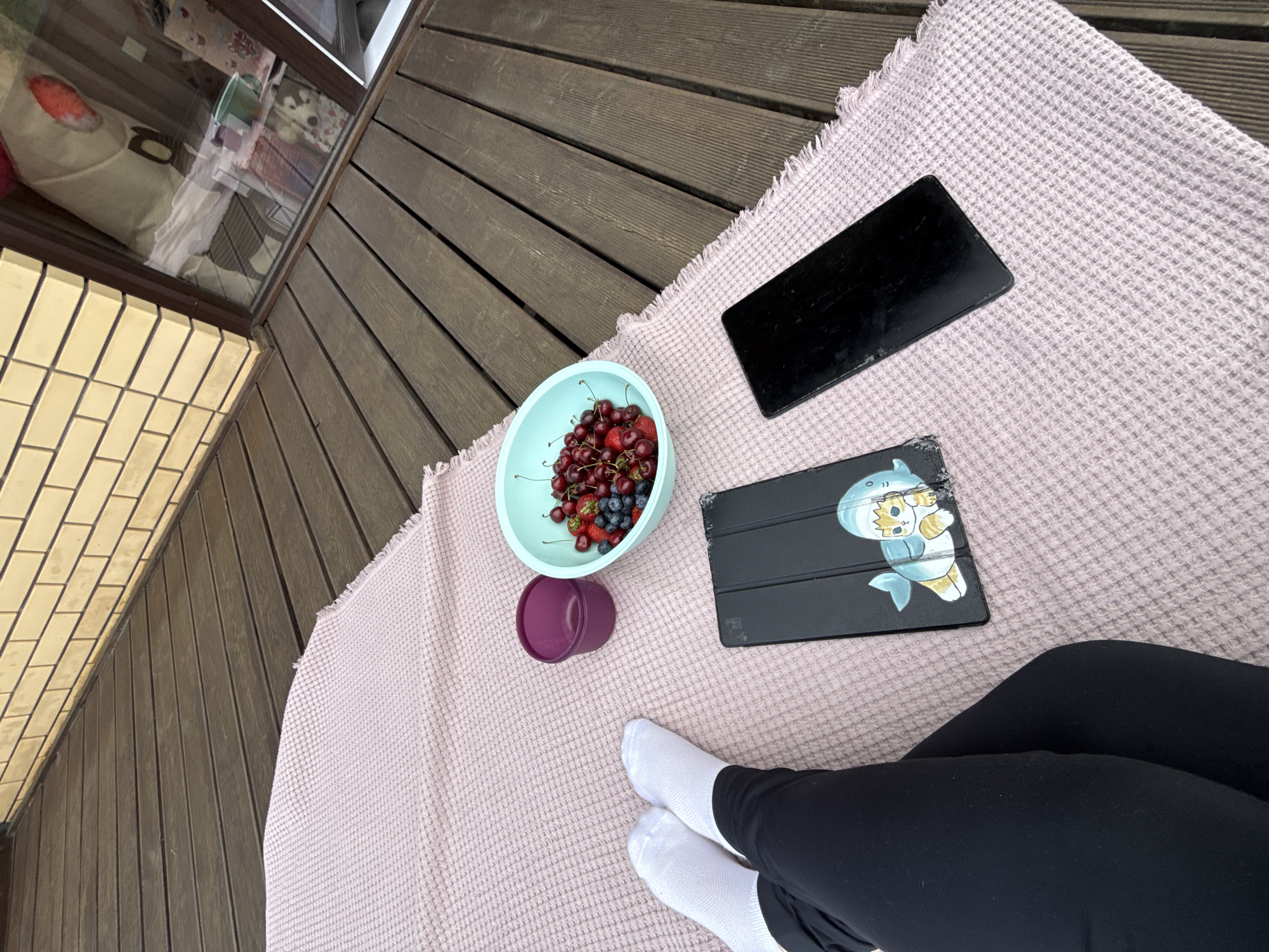 This image captures a first-person, slightly elevated view of a person relaxing on a wooden deck or balcony in Molodezhnyi, Russia. The person's lower legs, dressed in black pants or leggings and white socks, are visible in the lower right foreground, resting on a light pink, waffle-textured blanket.
On the blanket, a light blue bowl is prominently featured, filled with a vibrant mix of fresh fruits, including red cherries, red strawberries, and blue blueberries. Adjacent to the fruit bowl is a dark purple cup. Two black electronic devices also lie on the blanket; one is a larger tablet with its screen turned off or facing downwards, and the other is a smaller device, possibly a phone or e-reader, encased in a black cover adorned with a playful cartoon graphic of a shark holding a cat.
The wooden floor of the deck, constructed from dark, horizontally laid planks, stretches into the background. To the left, a section of a light yellow brick wall is visible, while a window reflects an interior space, showing what appears to be bags and other items. The natural light suggests it is daytime, creating a relaxed and leisurely atmosphere for enjoying snacks and possibly engaging with digital content. No discernible text is visible within the image.