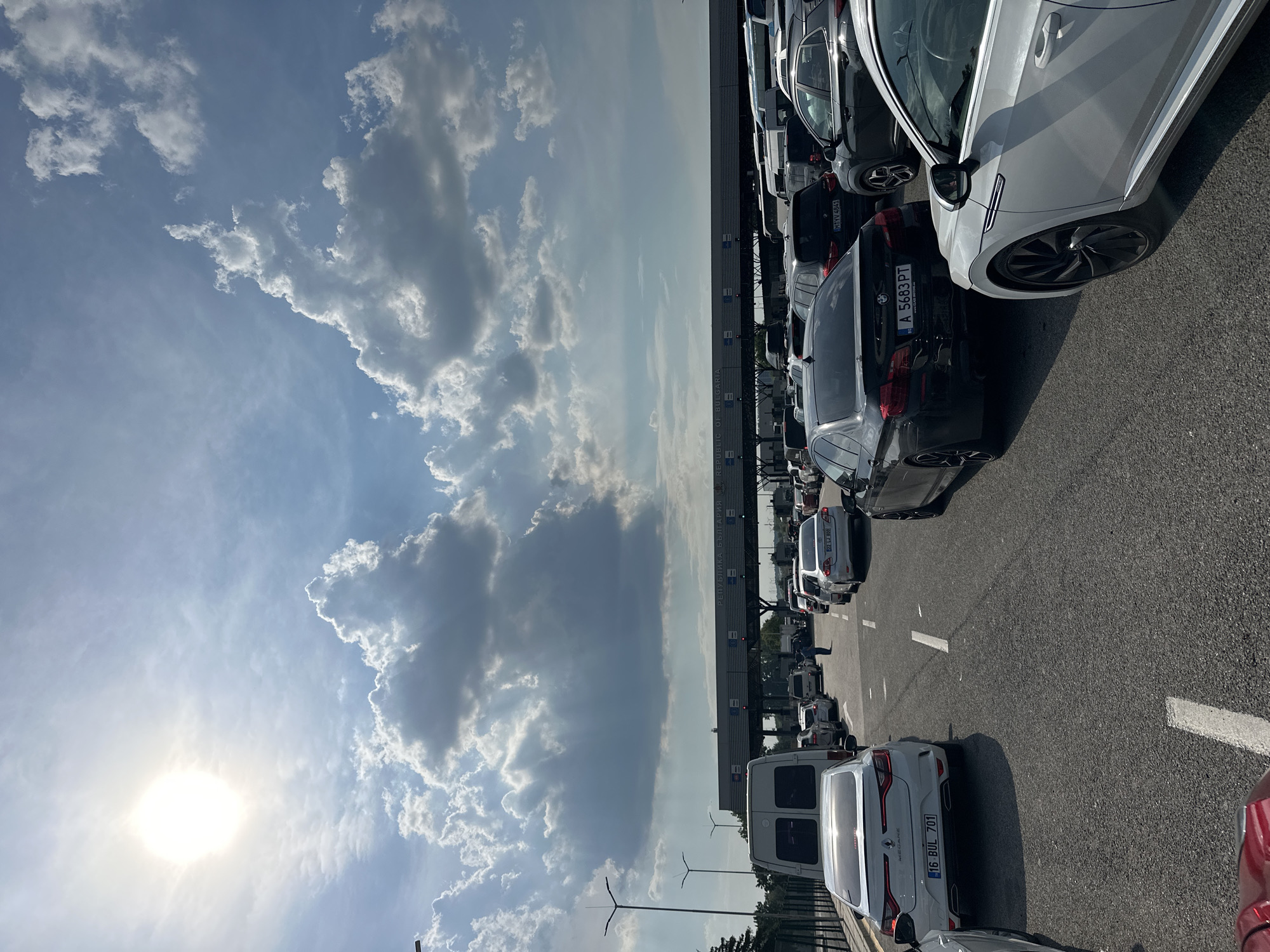 The image displays a bustling daytime scene at the Kapitan Andreevo border crossing into Bulgaria, characterized by a long queue of vehicles under a partly cloudy sky. The perspective is from inside a red vehicle situated in one of the multi-lane approaches to the border checkpoint.
The upper portion of the image shows a bright blue sky dominated by scattered cumulus clouds, some casting visible crepuscular rays. The sun is prominent in the upper left, emitting a strong glare and lens flare, indicative of a high sun angle.
The lower half captures the asphalt road, segmenting into multiple lanes packed with queued cars. A large, grey overhead structure spans across these lanes, featuring "РЕПУБЛИКА БЪЛГАРИЯ" and "REPUBLIC OF BULGARIA" in white lettering, confirming the border entry point. Below this main sign, several smaller panels or electronic displays are visible. On the left side of the road, two streetlights on poles and a metal fence run parallel to the traffic.
Vehicles of various makes, models, and colors are densely packed in the queues. In the leftmost visible lane, a white Renault Megane sedan (Turkish plate: 16 BUL 701) is followed by a white commercial van. In the central lane, a black BMW 5-series sedan (Bulgarian plate: A 5683 PT) is in the immediate foreground, followed by a dark SUV (German plate: M:VV 4841). Further down this lane, a single individual stands beside a vehicle. On the right, partially visible, is a white Volkswagen Arteon, followed by a dark SUV and a white sedan (Turkish plate: 32 EJ 800). Numerous other cars and vans extend into the background across all lanes, suggesting significant traffic congestion.
The overall environment is an open-air border facility, with minimal surrounding foliage immediately adjacent to the road, beyond the fence. The primary activity depicted is the slow, stationary queueing of vehicles awaiting processing at the border.