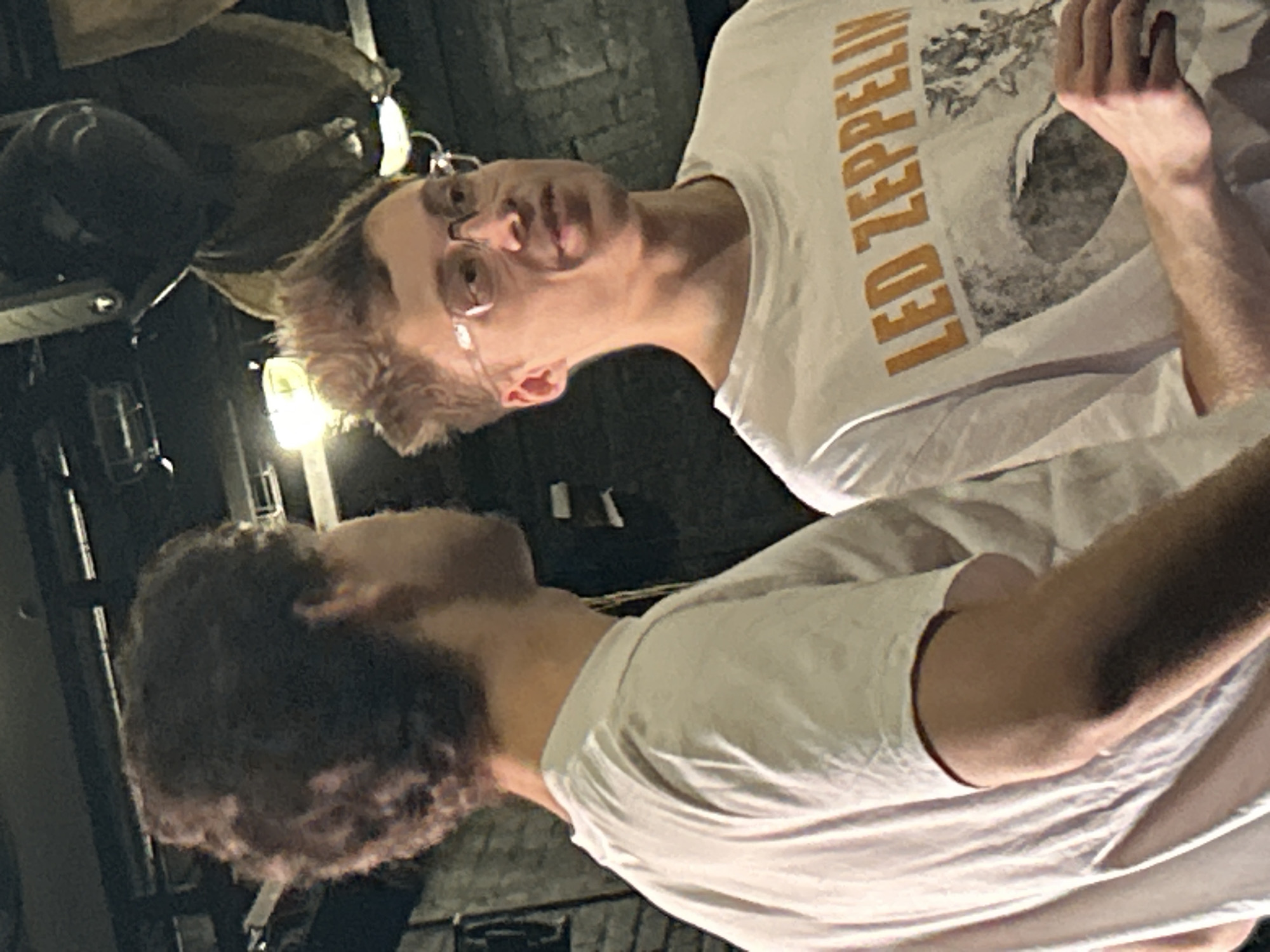 The media file shows two people in a dimly lit, indoor setting. The person in the foreground, seen from the back and side, has curly dark hair and is wearing a white t-shirt. The person in the background is seen from the front and side, with blonde hair and glasses. They are also wearing a white t-shirt with "LED ZEPPELIN" printed in orange text. The image appears to be taken at night, and the lighting suggests an indoor location such as a club or a home. The mood is casual and relaxed, with both individuals appearing to be engaged in conversation or looking at something together. There are no visible weather cues, and the location is not identifiable from the provided image.