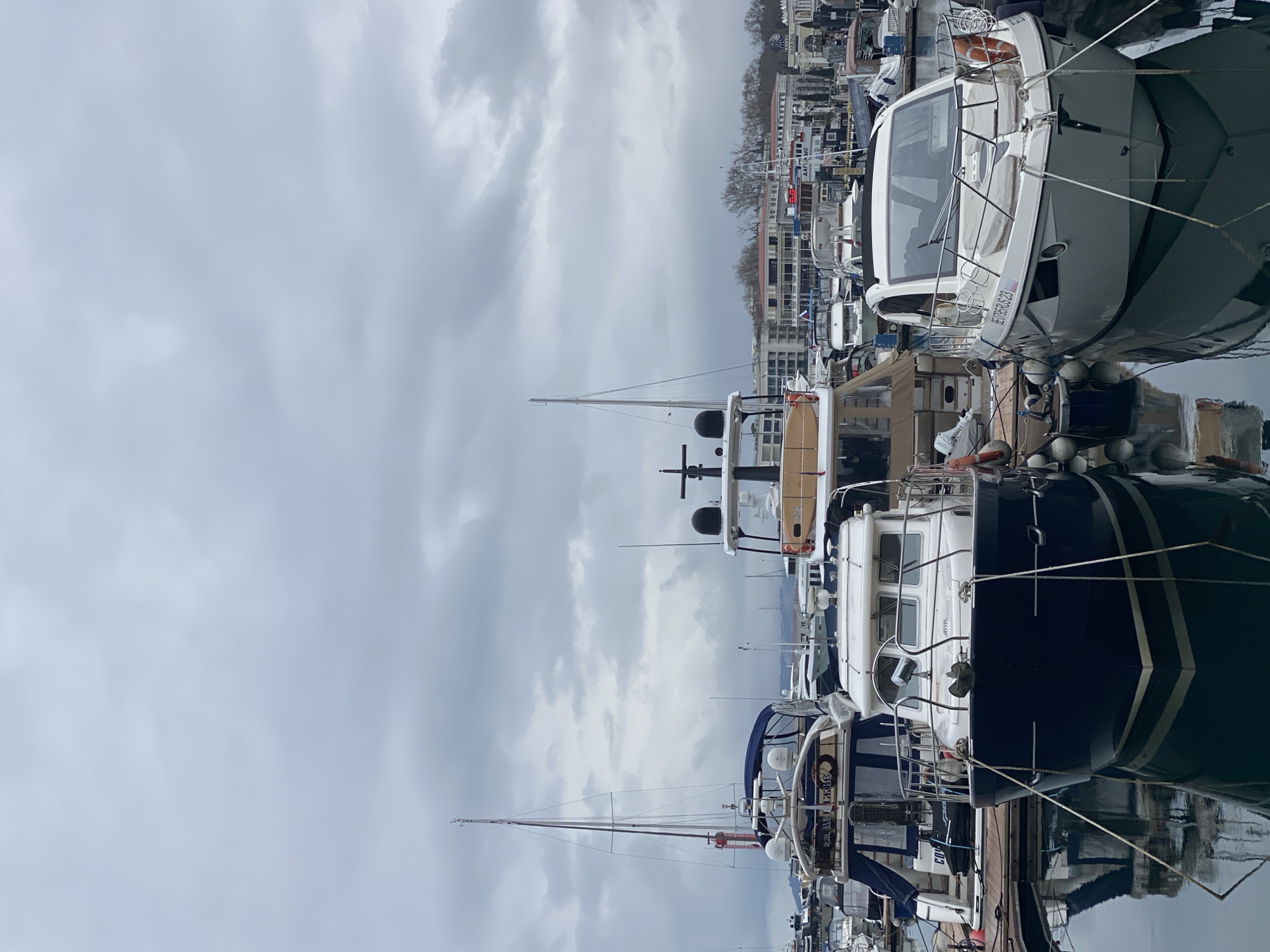 The image depicts a marina in Sochi, Russia, with multiple boats of varying sizes docked close to each other. The sky is overcast with heavy clouds, and reflections of the boats and the sky can be seen in the water. The buildings of Sochi are visible in the background. The focus is on the boats in the foreground, which are large and well-maintained.