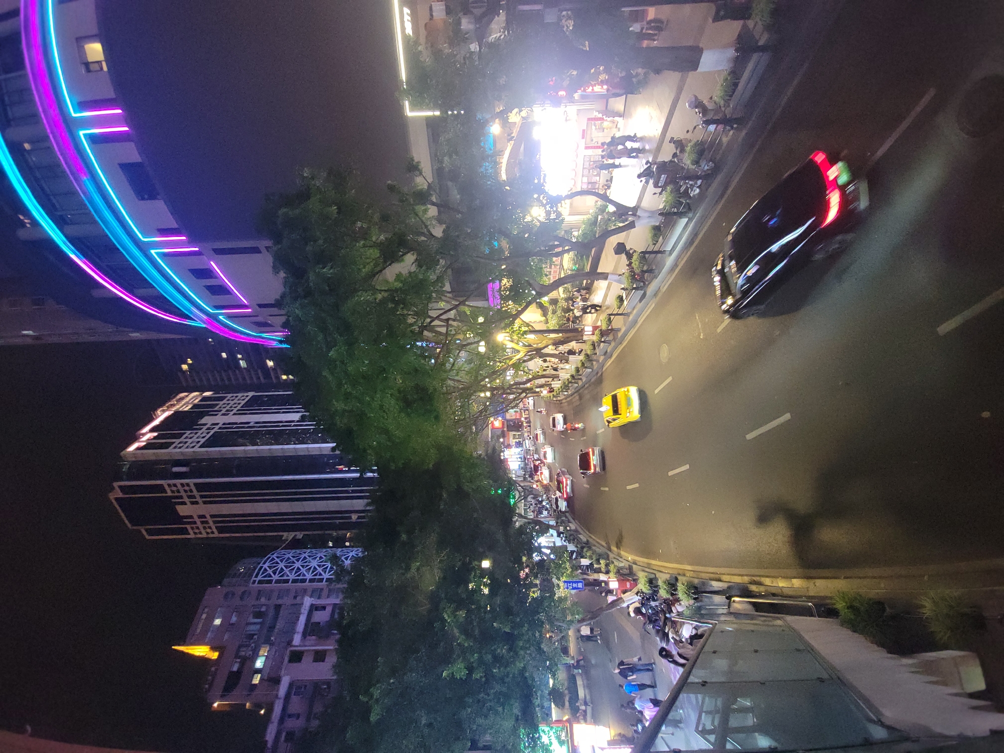 This image captures a vibrant, illuminated street scene at night, viewed from a high vantage point overlooking a bustling urban area, consistent with Yuzhong District, China. The scene depicts a multi-lane road curving through the city, flanked by tall buildings and tree-lined sidewalks.
The prominent feature on the left is a large, modern building adorned with striking, curved neon lights in vivid blue and magenta hues, illuminating its facade. Other tall buildings, some with intricate architectural patterns and illuminated windows, rise against the dark night sky. A dense line of trees runs along the street, their foliage glowing under the numerous streetlights, which also cast long shadows across the road and sidewalks.
The street is active with vehicles; a bright yellow taxi is visible, along with a black car showing distinctive red taillights, suggesting movement. Motorcycles and scooters are parked along the sidewalks, where numerous people are seen walking, standing, and congregating. Some areas along the sidewalk appear to be brightly lit commercial spaces or outdoor dining establishments, bustling with activity.
The time of day is clearly night, evidenced by the dark sky and the reliance on artificial lighting throughout the scene. The atmosphere is lively and energetic, characteristic of a major city. A blue street sign displaying Chinese characters is visible. In the immediate foreground, a glass-covered structure, possibly part of a building entrance or pedestrian overpass, frames the bottom of the image. The overall impression is a dynamic and thriving urban environment after dark.
