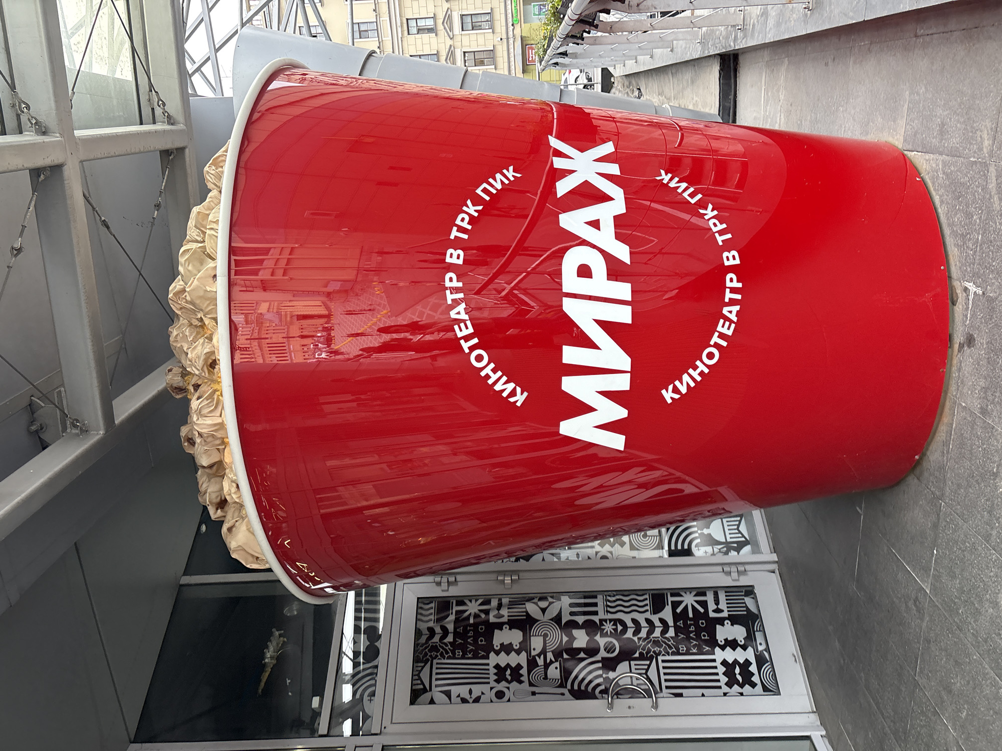 This is an outdoor photograph of a large, red popcorn bucket, advertising the "Mirazh" cinema at the PIK shopping mall. The bucket is positioned at the entrance of a building. The bucket has the words "MIRAZH" in large white letters, surrounded by the Russian text "КИНОТЕАТР В ТРК ПИК," which translates to "Cinema in TRK PIK." Inside the bucket, faux popcorn protrudes from the top. The scene is captured during daylight, with reflections visible on the bucket's glossy surface. The location is Saint Petersburg, Russia.