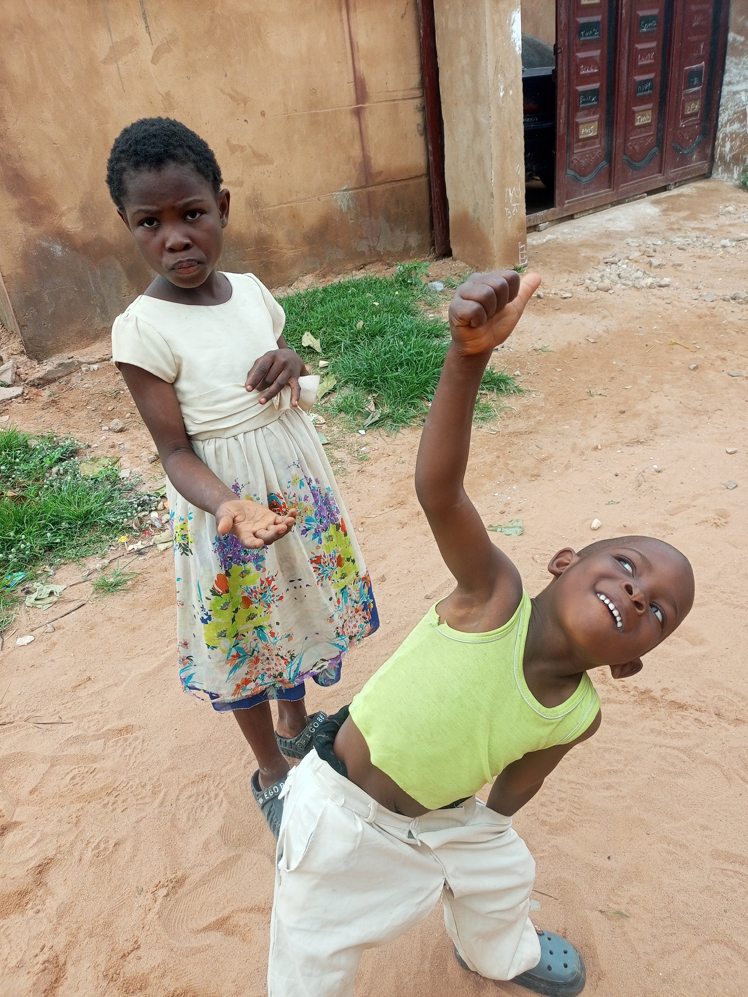 Two children are standing on a dirt ground in front of a building with a closed metal gate. The child on the left, a young girl, wears a light-colored dress with a floral pattern and stands with her left arm slightly extended, her hand open as if offering something. The child on the right, a young boy, wears a lime green tank top and light-colored pants. He is leaning back with his right arm raised, fist clenched and thumb up, and has a wide smile. The environment appears to be a residential area in Tempure, Nigeria, characterized by dirt paths, some sparse grass, and simple building structures.