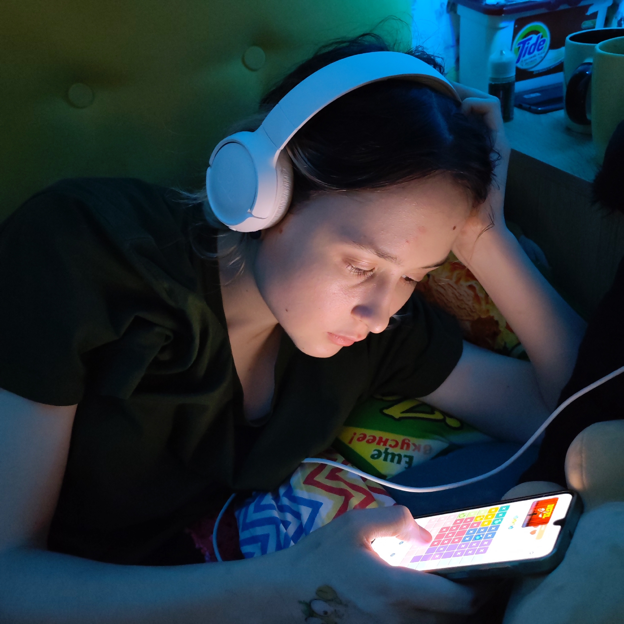 A young woman is lying on a green-colored couch. She is wearing white headphones and a dark green t-shirt. She is looking at a smartphone in her hands, her finger touching the screen which displays a colorful puzzle game. A white charging cable is connected to the phone. The background includes a pillow with a multi-colored zig-zag pattern, a green bag of "Eщё вкуснее!" chips, and items on a table nearby. The word "Tide" is visible on one of these items. The location is Chaikovsky, Russia.
