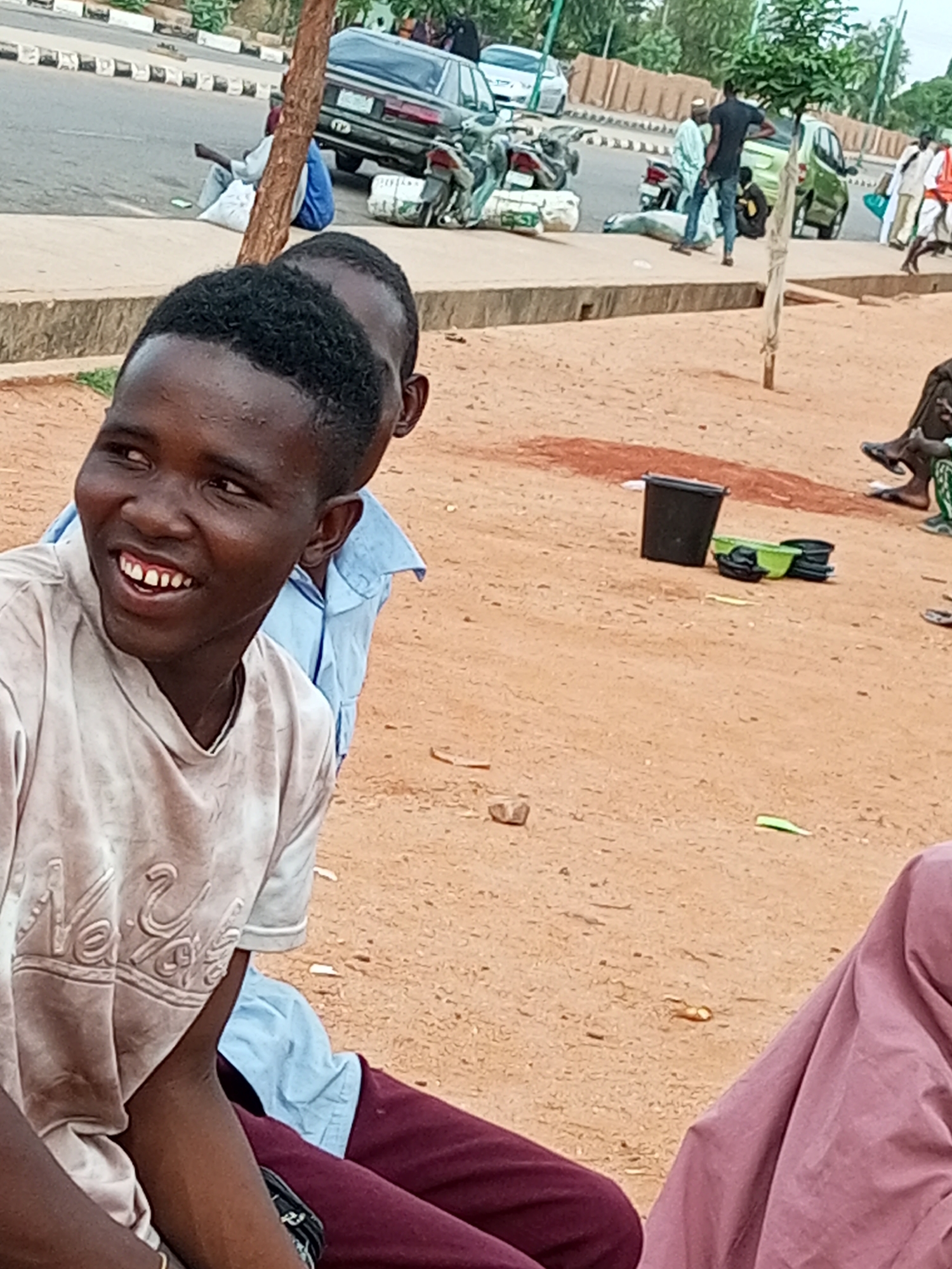 The image depicts a street scene in Gombe, Nigeria, with several individuals present. In the foreground, a young man with dark skin and short, black hair is smiling and looking to his left. He is wearing a light-colored, possibly dirt-stained t-shirt with a raised design on the front. Partially visible behind him is another person wearing a light blue shirt.
In the background, a road is visible with several vehicles, including a dark sedan, a light-colored sedan, and a green SUV. Several people are standing or sitting near the road, some appearing to be engaged in conversations or tending to goods. A tree trunk with a white ribbon tied around it stands in the middle ground. The ground is covered in sand and some debris. On the sandy ground, a black bucket and some green plastic containers are visible. The overall environment appears to be an urban setting, possibly a market or a busy roadside area.