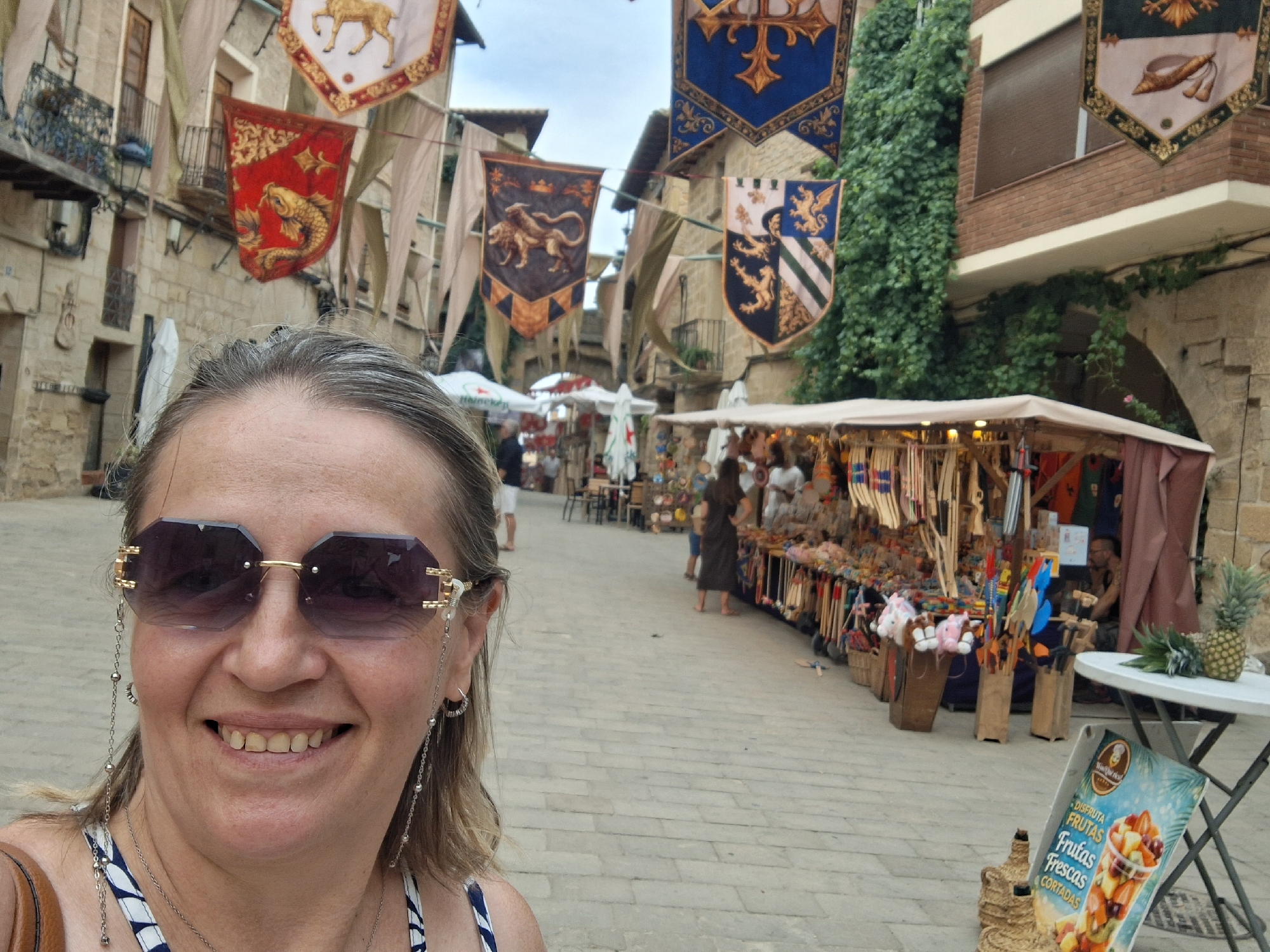 In La Fresneda / la Freixneda, Spain, a woman wearing large, dark sunglasses with decorative chains attached to the frames is taking a selfie. She is positioned in the foreground of the image, smiling directly at the camera. Her hair is blonde and styled loosely. She is wearing a white and blue striped garment. Behind her, a cobblestone street is lined with market stalls adorned with medieval-style banners. One banner features a golden stag, another a red background with a golden fish, and others display coats of arms with lions and eagles. The stalls sell various goods, including wooden crafts, textiles, and food items. A pineapple sits on a white table to the right, next to a sign advertising "Disfruta Frutas Frescas Cortadas" (Enjoy Fresh Cut Fruits). People are browsing the stalls in the background, and buildings with stone facades are visible along the street. The sky is overcast.
