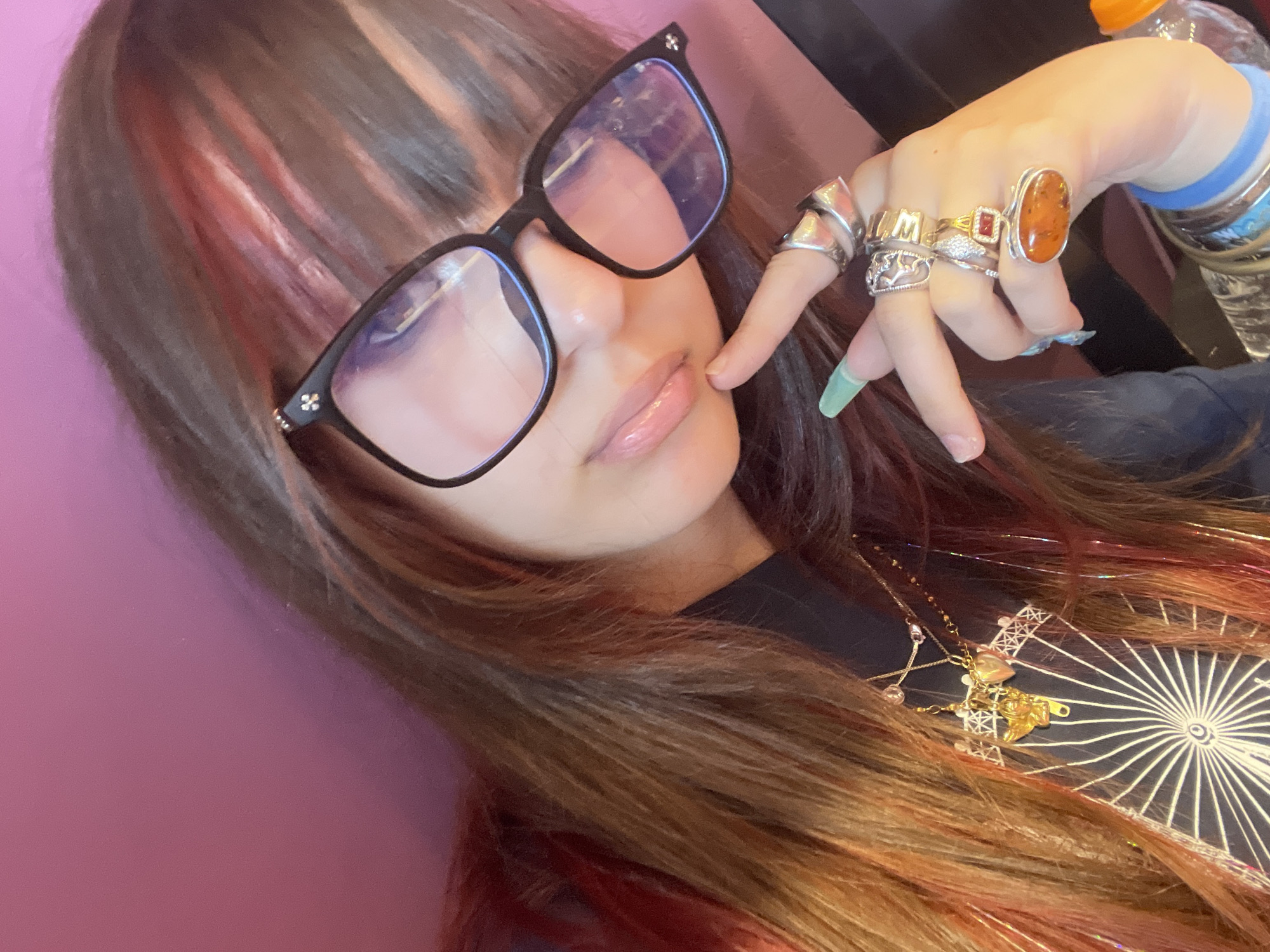 The image shows a close-up, waist-up view of a young woman with long, dark brown hair with streaks of pink and blonde. She is wearing black-rimmed glasses and a black shirt. She is wearing several rings on her fingers, including one with a large amber stone and another with a red stone. Her fingernails are painted a light green color. She is also wearing multiple necklaces, one of which has a gold angel charm and a heart-shaped locket. Her left hand is raised, with her index finger resting on her chin. The background is a solid pink color. The scene is indoors, and there is no indication of its location in Raanana, Israel.